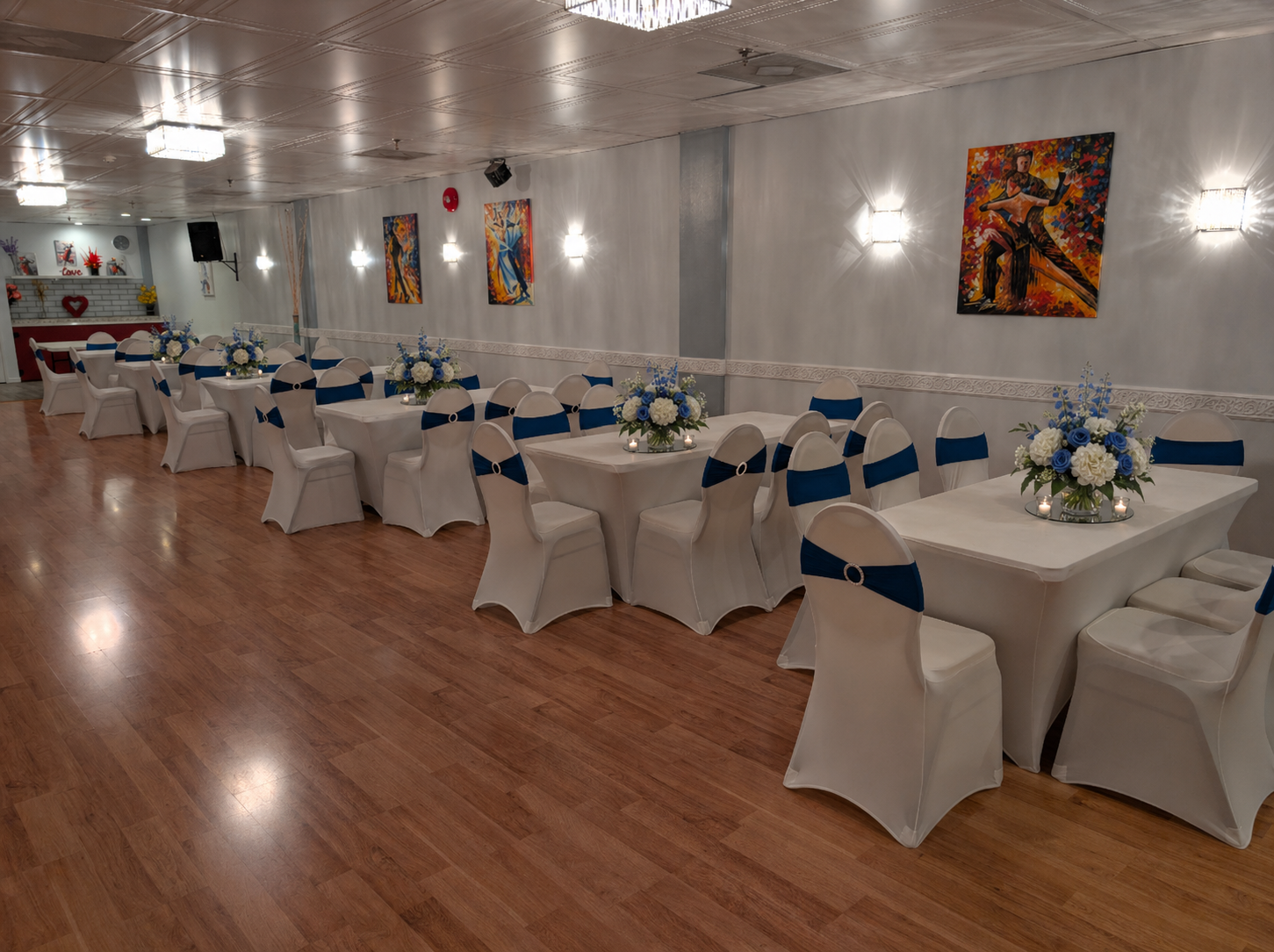 A banquet hall decorated for a formal event with white tablecloths, white chairs with blue sashes, and floral centerpieces with white and blue flowers. The color theme appears to be blue and white, with paintings on the wall and a wooden floor.