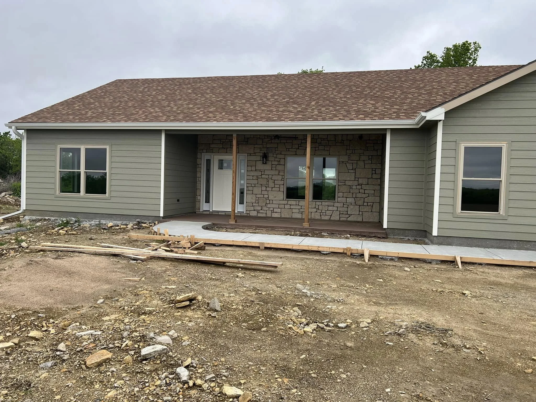 Front view of a house under construction with a brown shingled roof, beige siding, windows, and an unfinished porch area with construction materials on the ground.