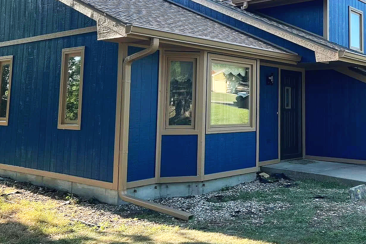 Exterior of a house with blue siding, beige window frames, a black front door, and a downspout installed by Gutter Solution LLC on the corner.