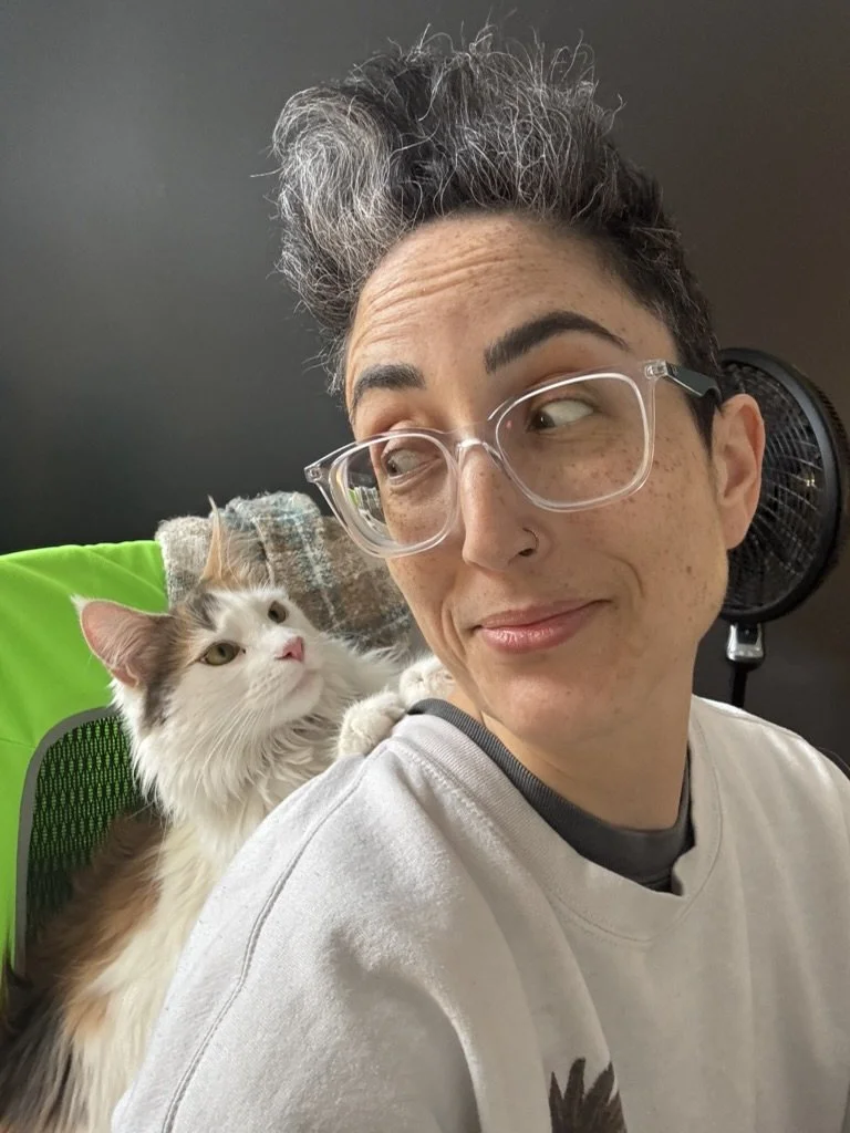 A person with short, gray and black hair, glasses, and a nose ring sitting in a room with a black fan in the background. A fluffy white and brown cat is perched on their shoulder. The person looks to the side with a smirk.