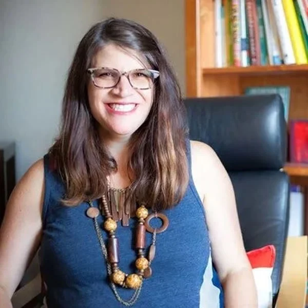 A woman with long brown hair, glasses, and a big smile sitting in a black office chair in front of a bookshelf with colorful books.