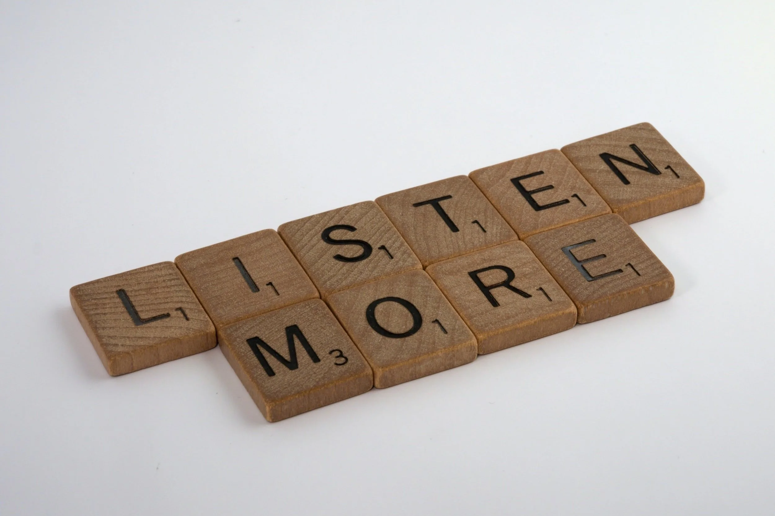 Wooden Scrabble tiles spelling out the word "LISTEN" on a white surface.