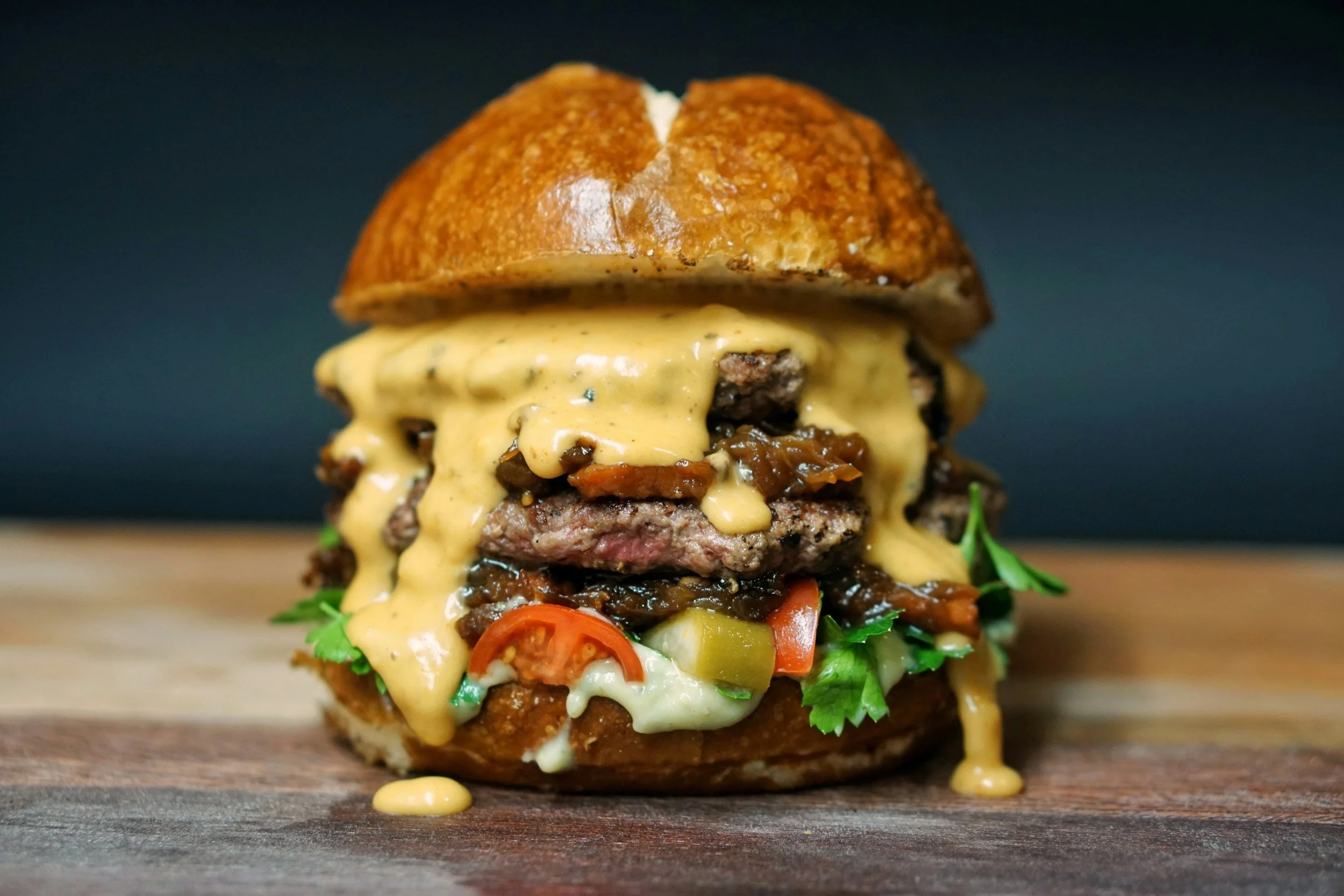 Close-up of a gourmet cheeseburger with melted cheese, beef patties, tomatoes, pickles, lettuce, and sauce in a brioche bun on a wooden surface.