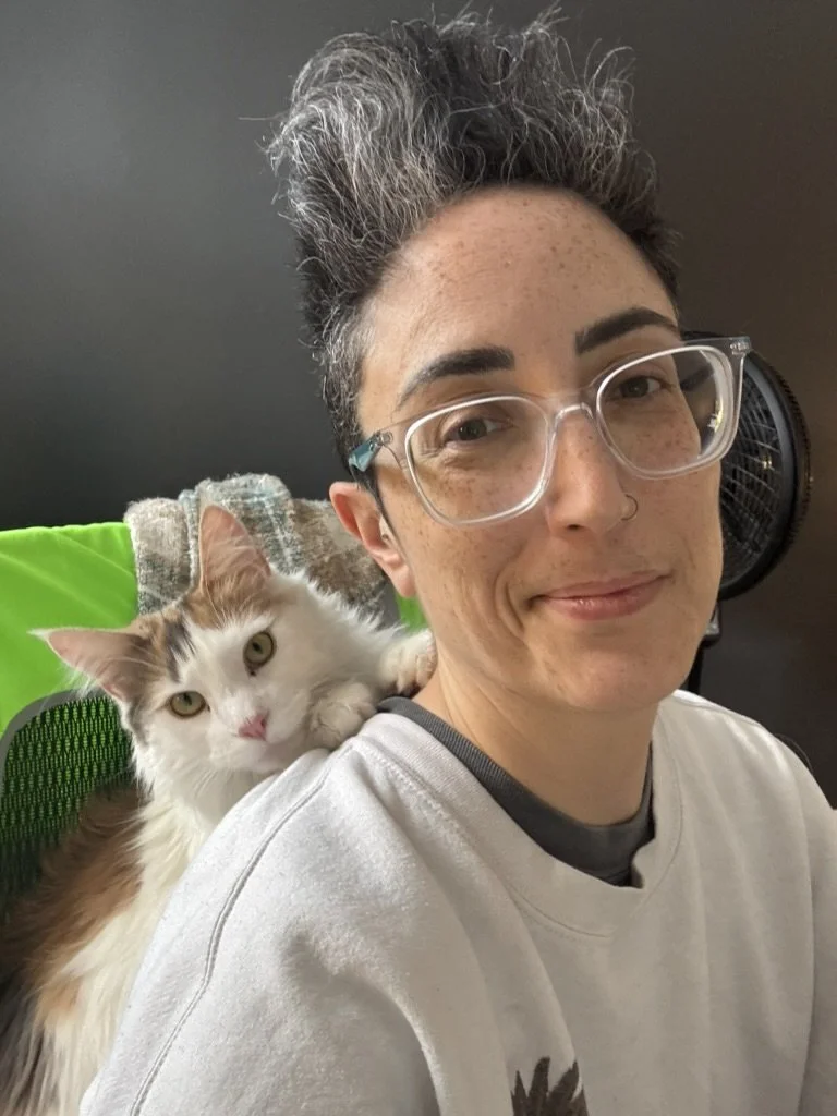 A person with short, gray and black hair and glasses, smiling, with a fluffy white and orange cat resting on their shoulder.