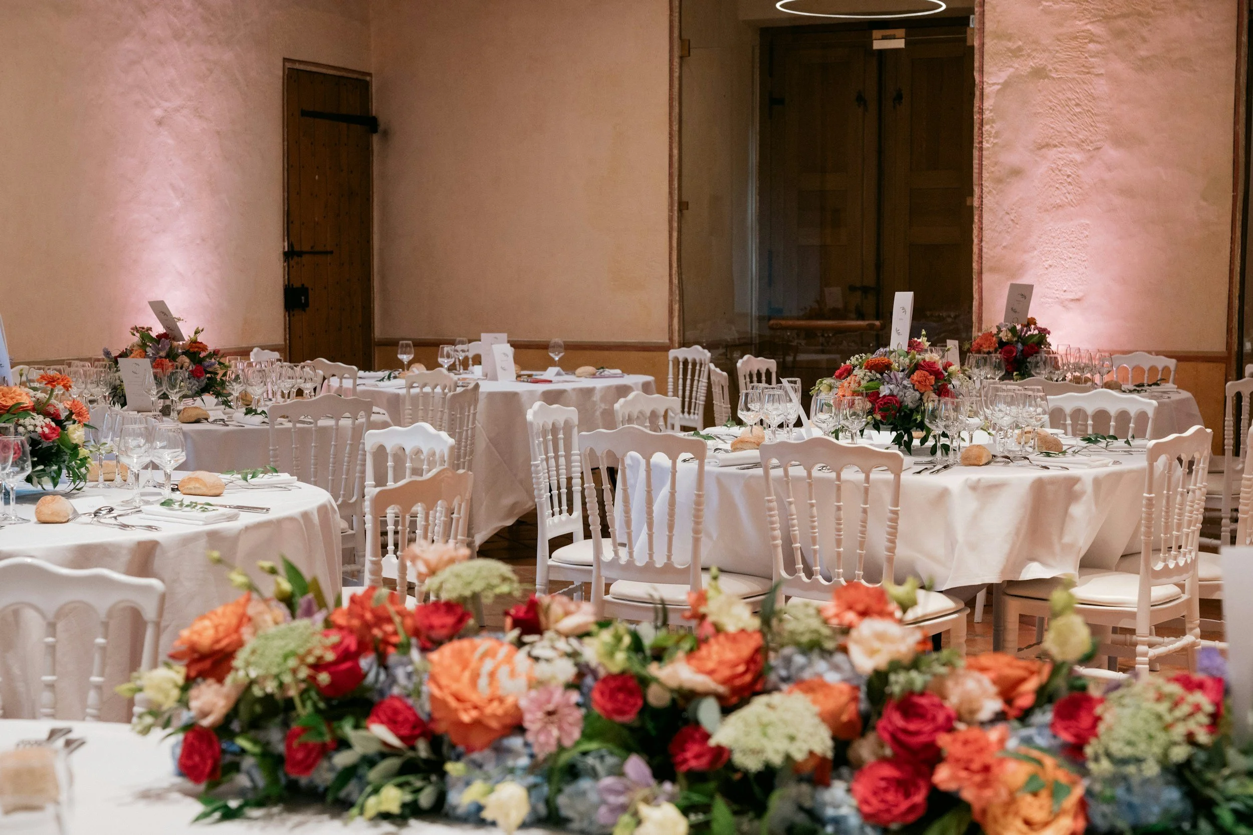 Wedding reception setup with tables decorated with floral centerpieces, glassware, and place settings in a banquet hall with beige walls and wooden doors.