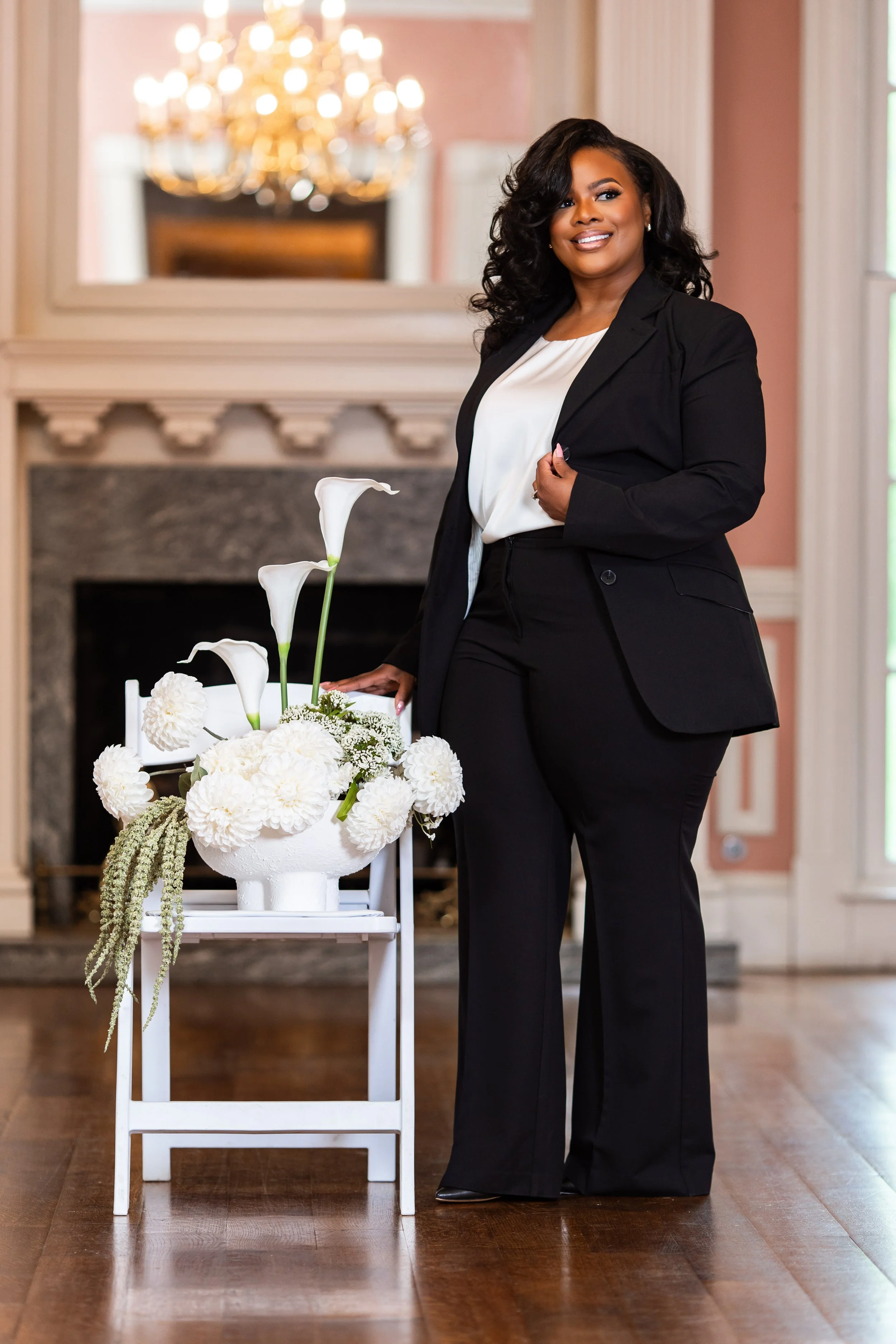 A woman in a black suit and white blouse standing next to a white floral arrangement in an elegant room with a chandelier and a fireplace.