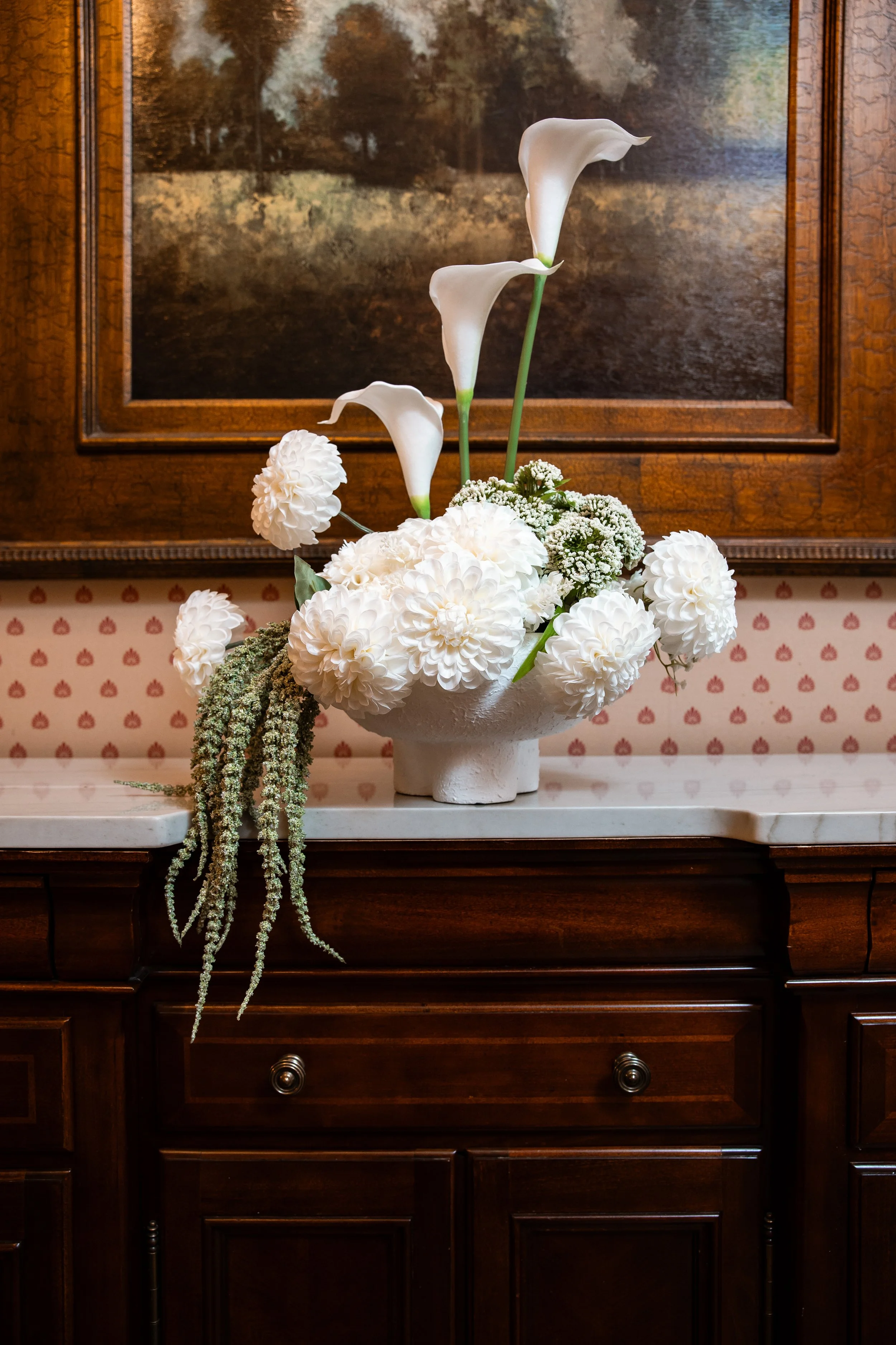 A floral arrangement with white calla lilies and white dahlias on a dark wooden sideboard against a framed landscape painting.