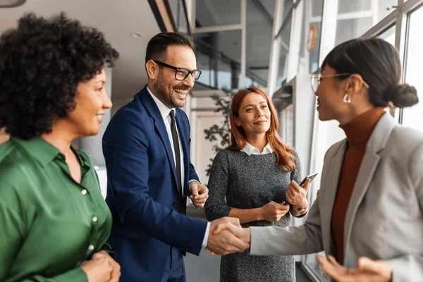A business woman in a gray suit shaking hands with a man in a blue suit, while two women look on in an office setting.