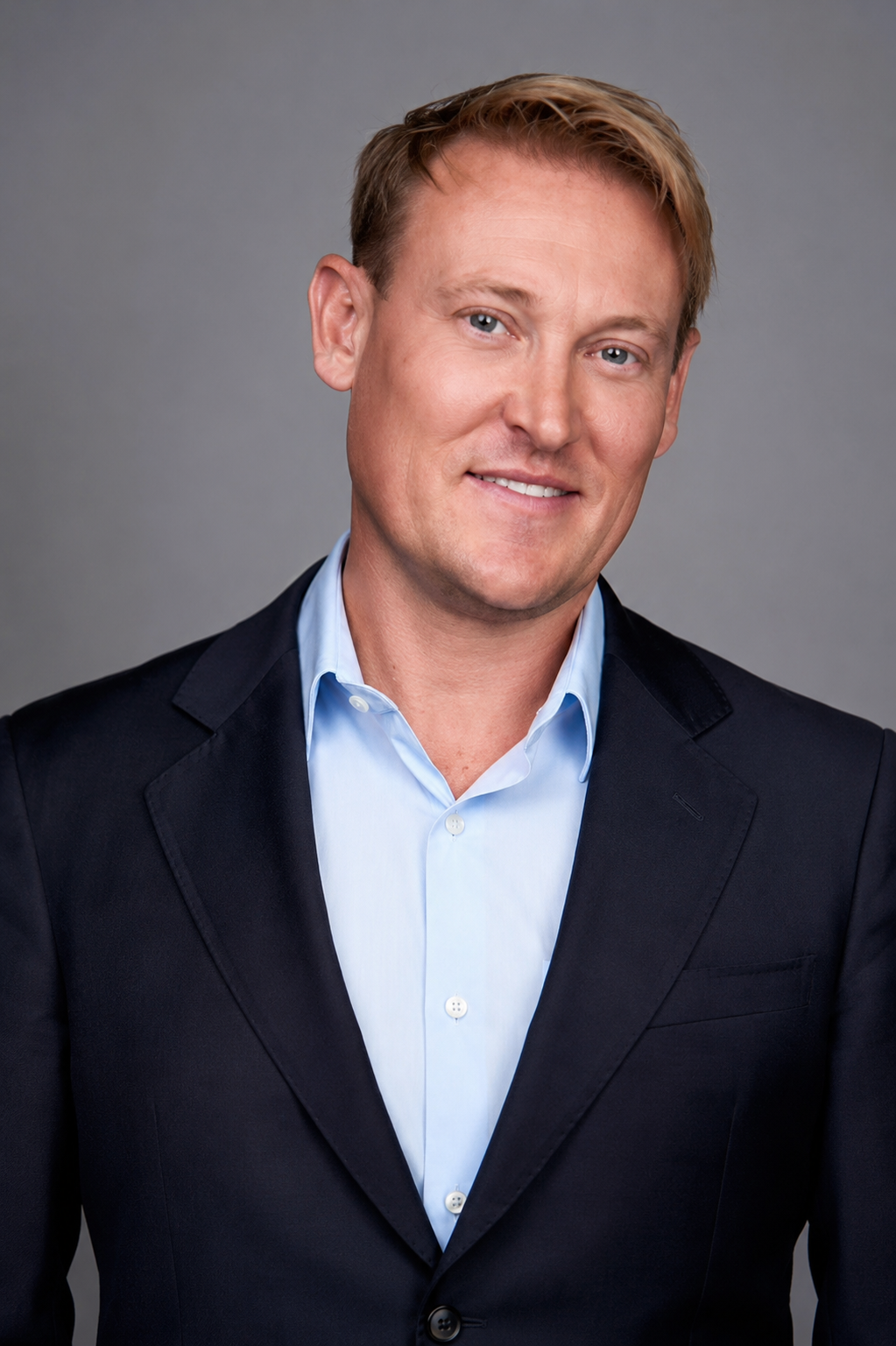A professional man in a dark suit and light blue shirt smiling against a gray background.