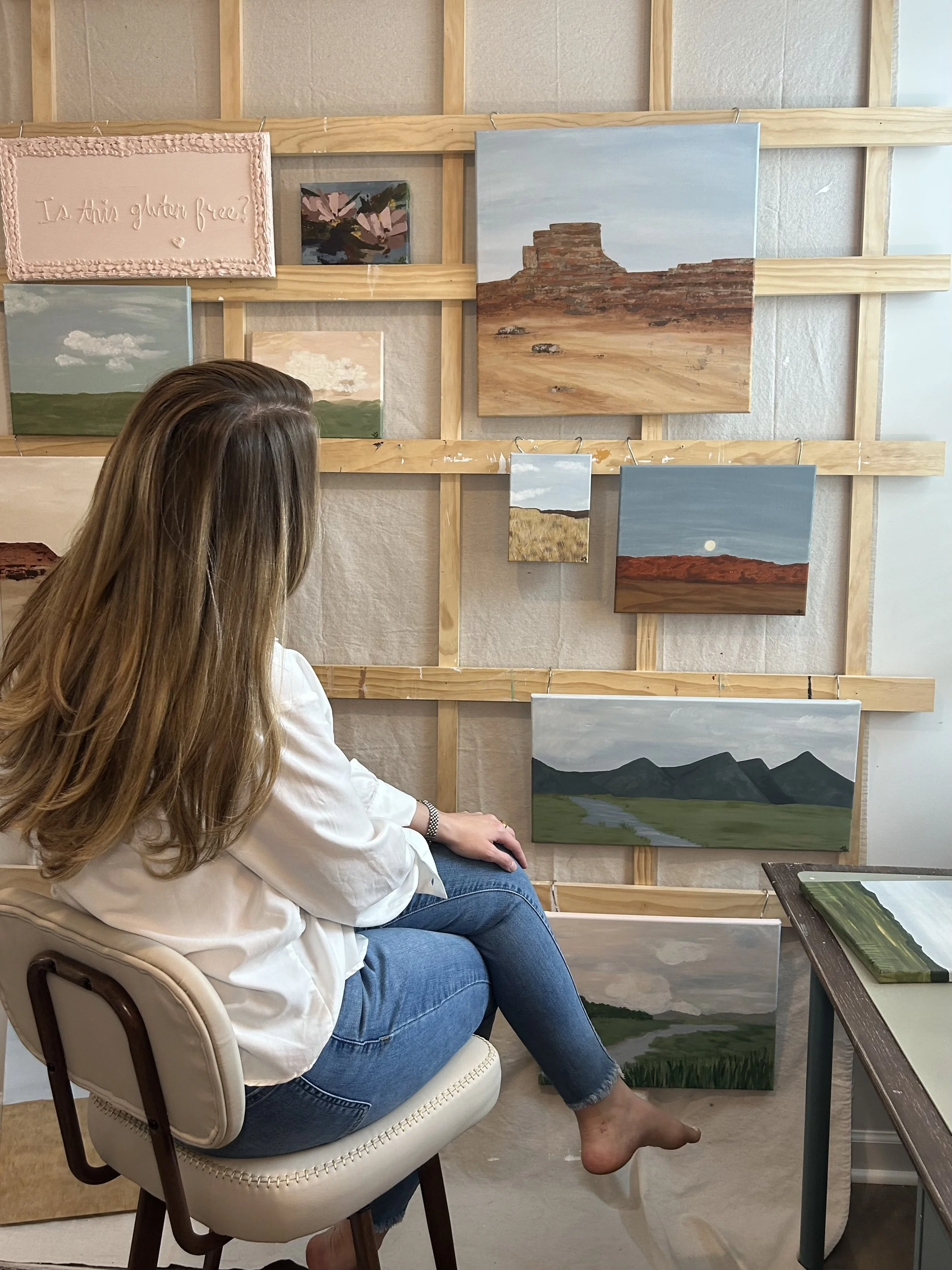 A woman with long hair wearing a white blouse and jeans, sitting on a chair in front of a display of landscape paintings on a wooden wall, inside an art gallery or studio.