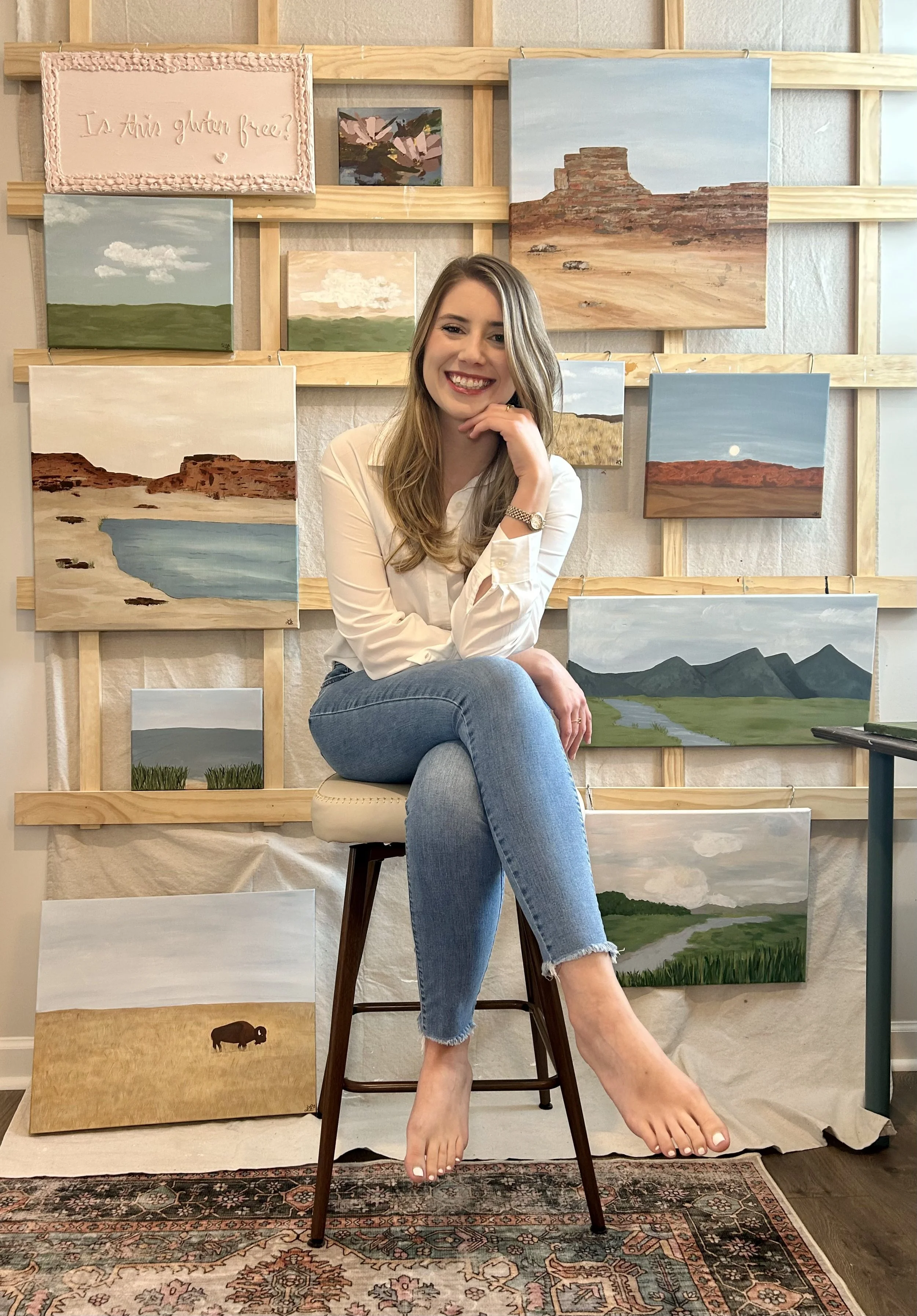 Young woman sitting on a stool, smiling, with multiple landscape paintings displayed on a wooden frame behind her, on a beige wall and carpeted floor.