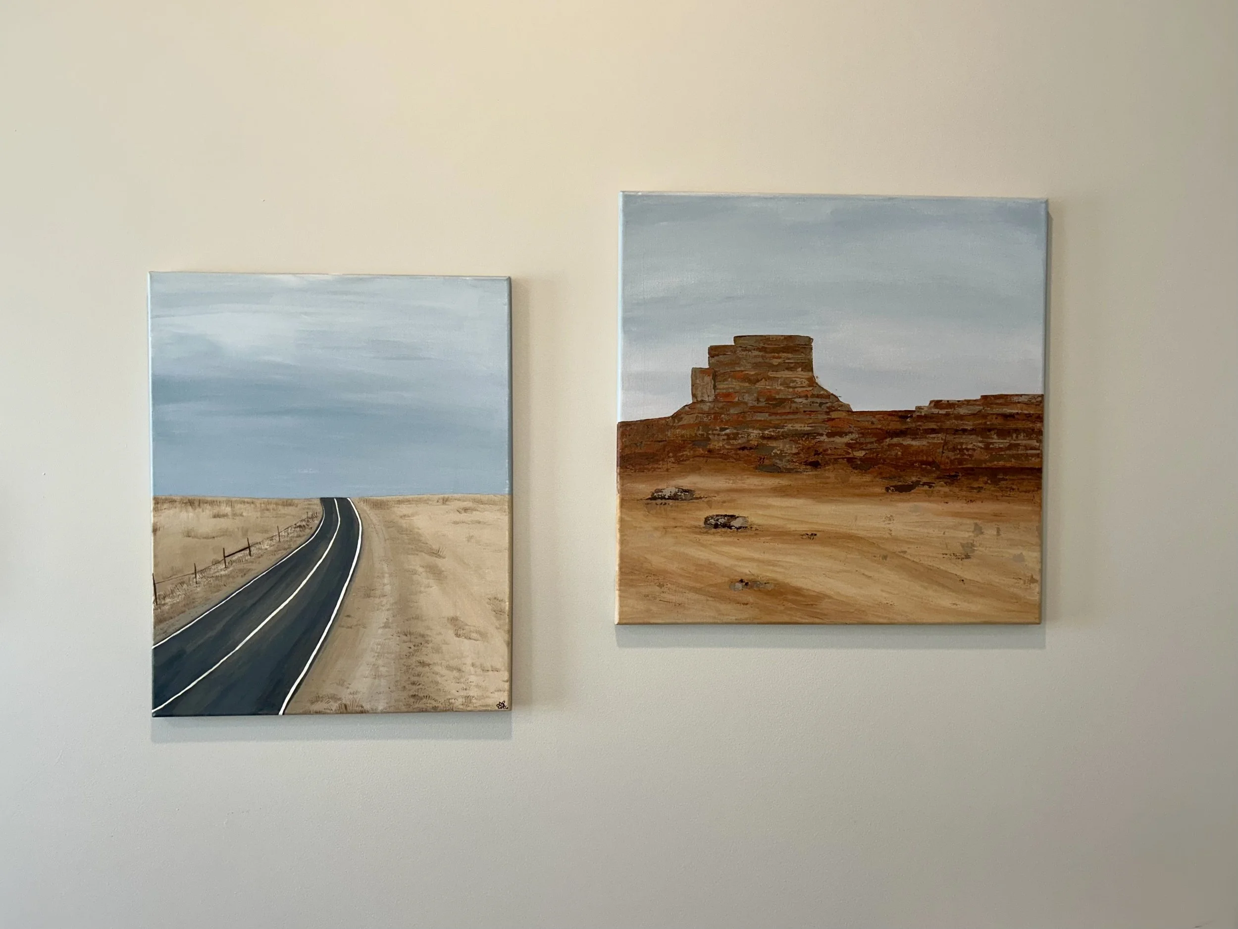 Two landscape paintings on a light-colored wall. The left painting depicts a highway scene with a road, dirt, and fence. The right painting features a desert landscape with a red rock formation and a cloudy sky.