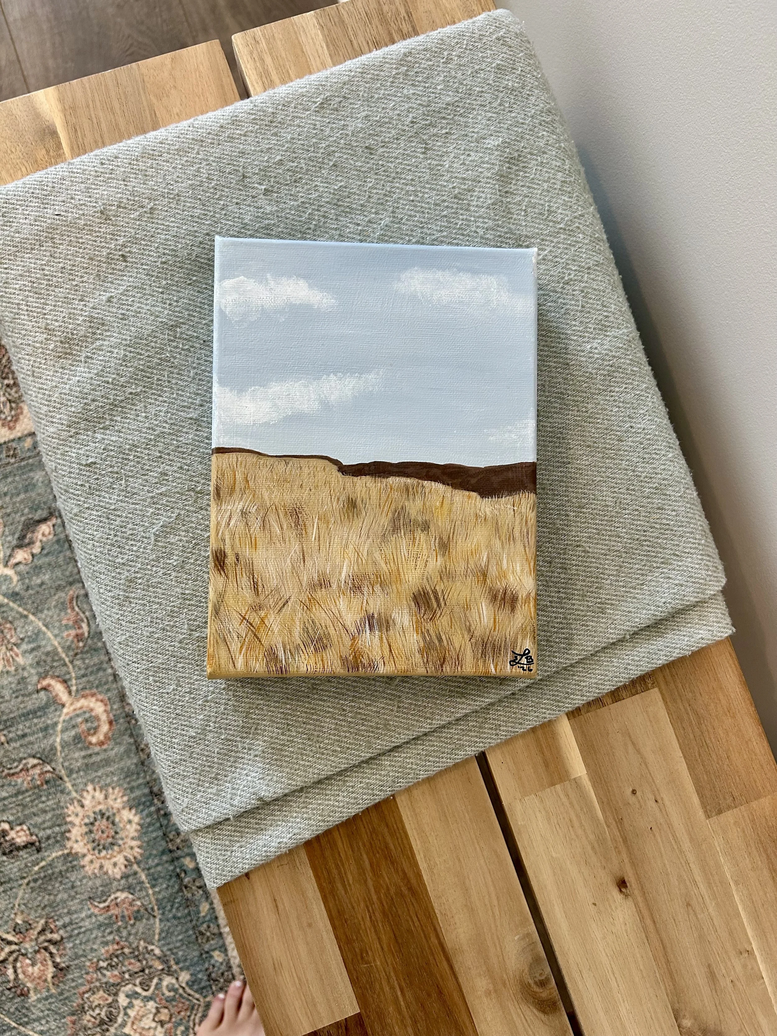 Small painting of a cloudy sky above a grassy field leaning against a fabric-covered surface, with a hand at the bottom of the image.