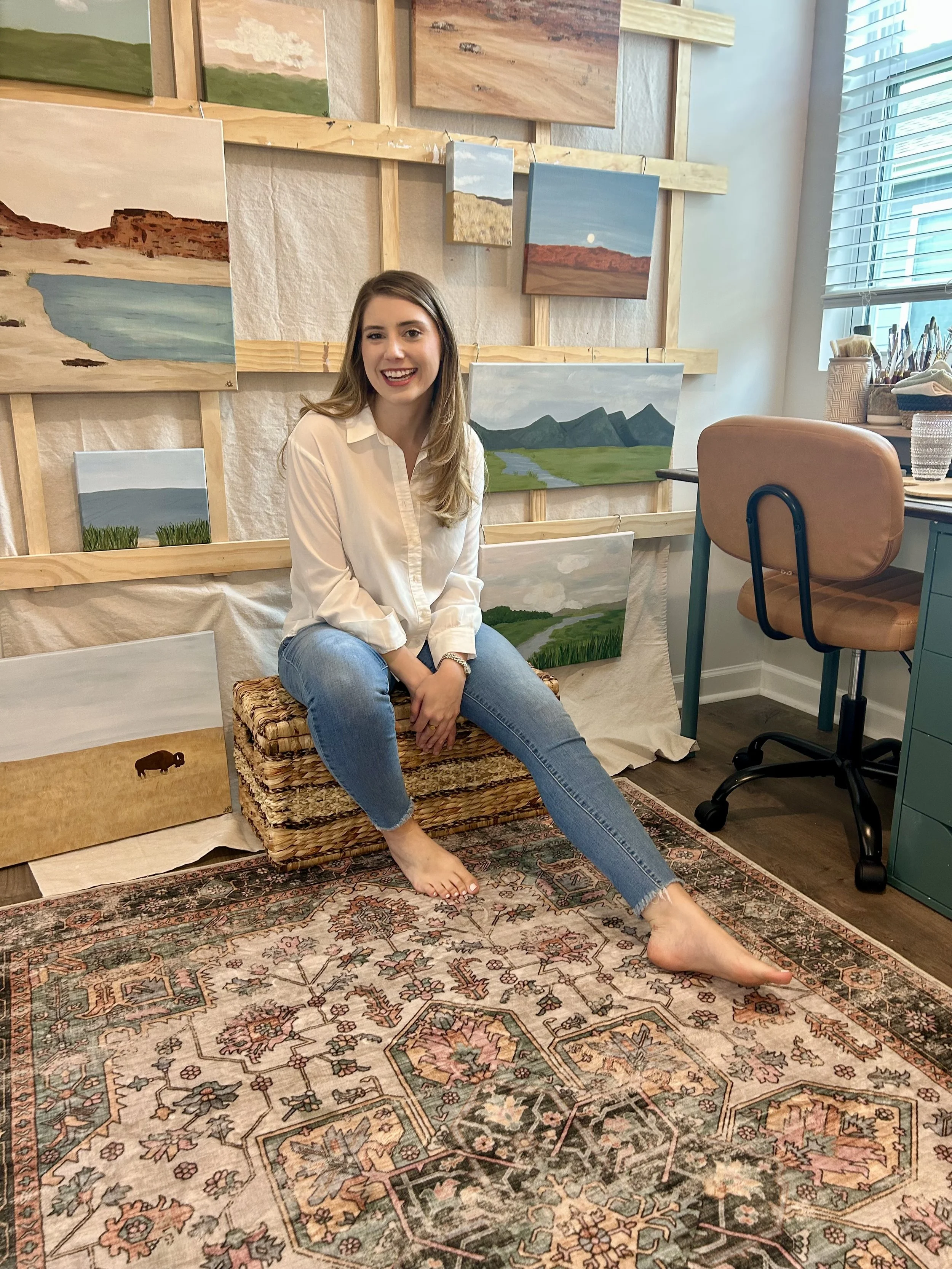 A woman sitting barefoot on a woven basket in an art studio, smiling at the camera, surrounded by landscape paintings on the wall and a patterned rug on the floor.