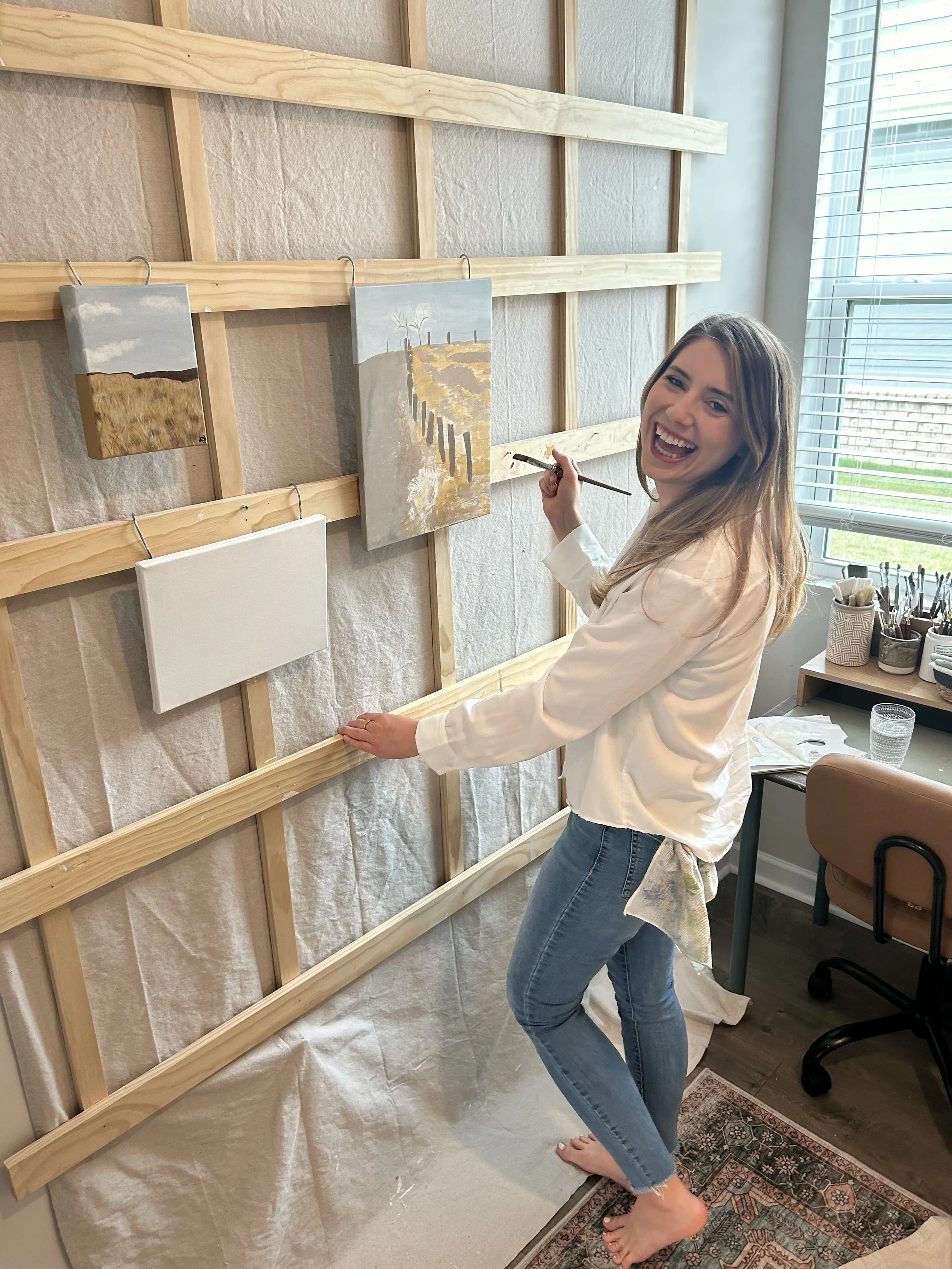A woman smiling and painting on a canvas mounted on a wooden frame in a well-lit room.