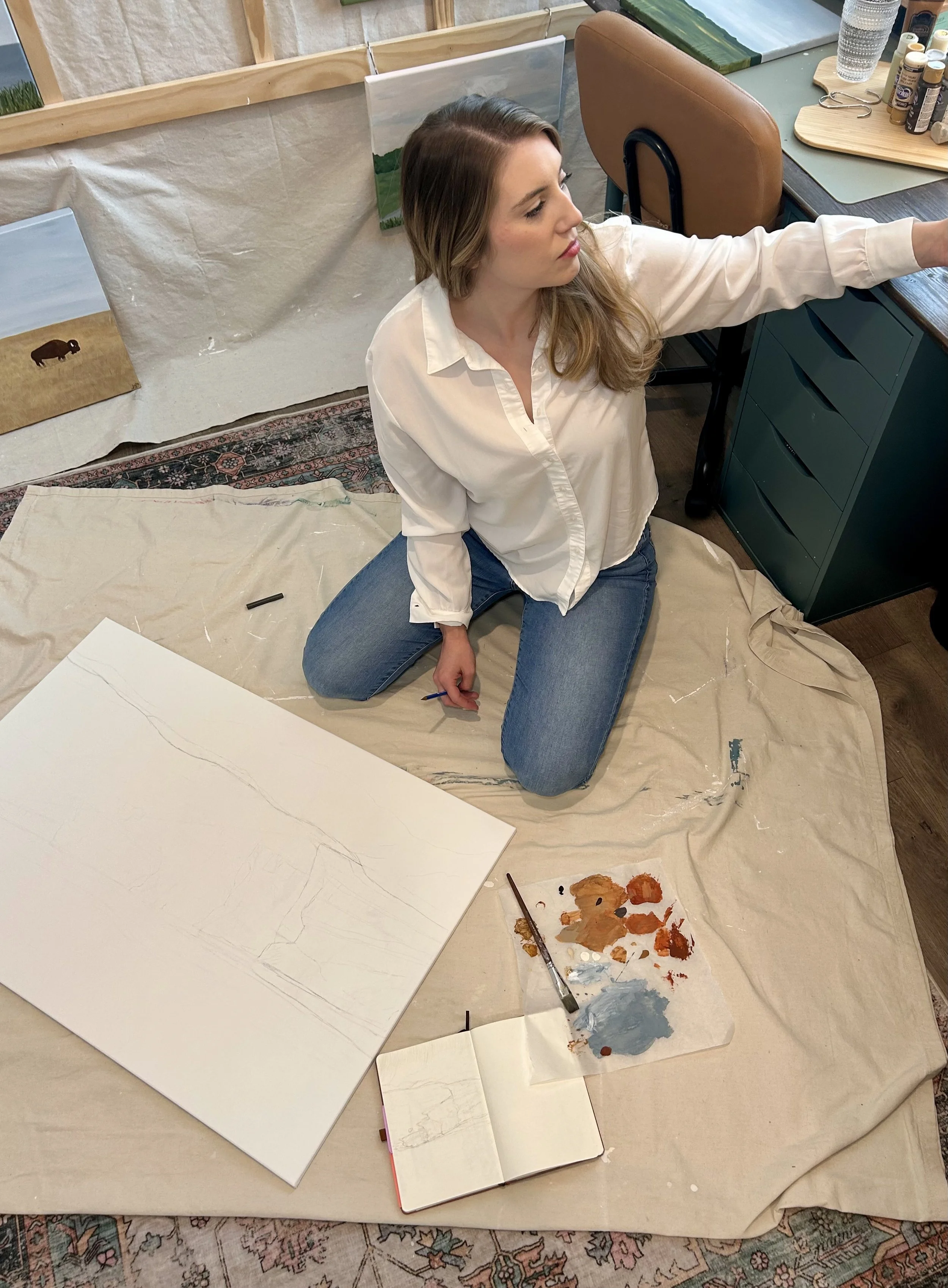 A woman with long, wavy brown hair, wearing a white blouse and jeans, kneeling on a beige cloth on the floor, holding a paintbrush in her right hand, in an art studio. In front of her are various paintings, including a blank canvas, an unfinished landscape, and a painting on a small notebook. There are art supplies nearby, including paints, a palette, and brushes. Behind her are more paintings leaning against the wall, and a table with additional art supplies and water glasses. The setting appears to be a creative workspace or art studio.