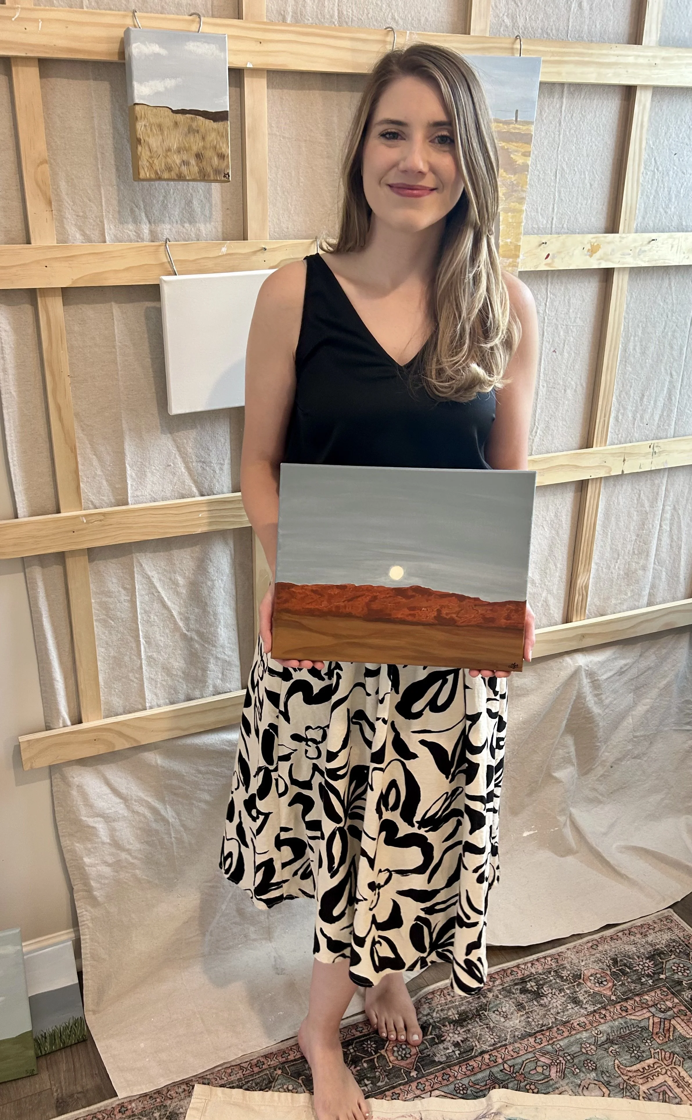 A woman holding a landscape painting of a desert with a moon in the sky, standing in front of wall art and wooden framework.