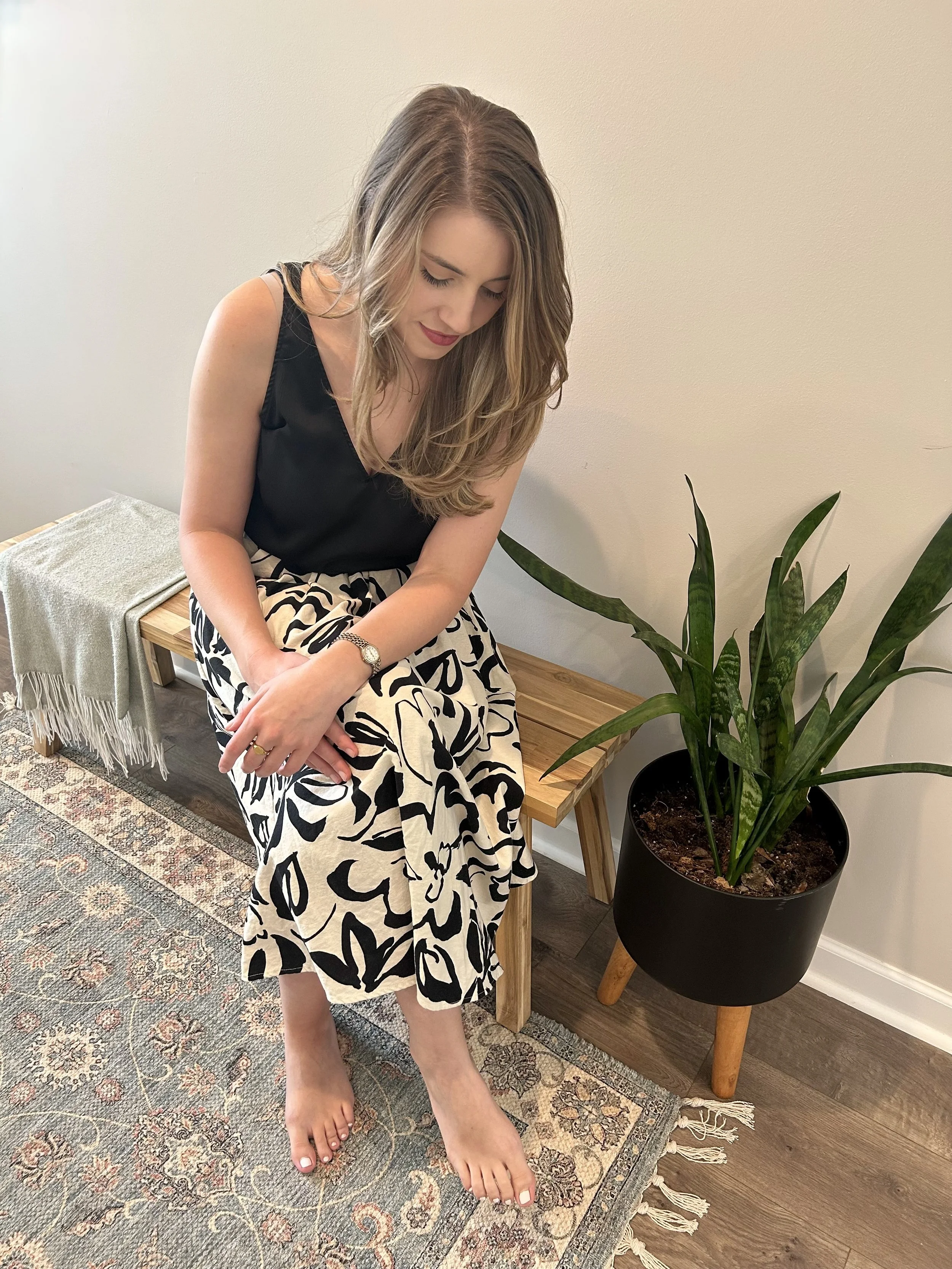 A woman with long, wavy hair sits on a wooden bench, looking down with her eyes closed. She is wearing a black sleeveless top and a black-and-white patterned skirt. She is barefoot, with her legs crossed at the ankles. Next to her is a large potted plant with long green leaves, and behind her is a beige wall. To her left, there is a small table with folded fabric, and the floor is covered with a patterned rug.