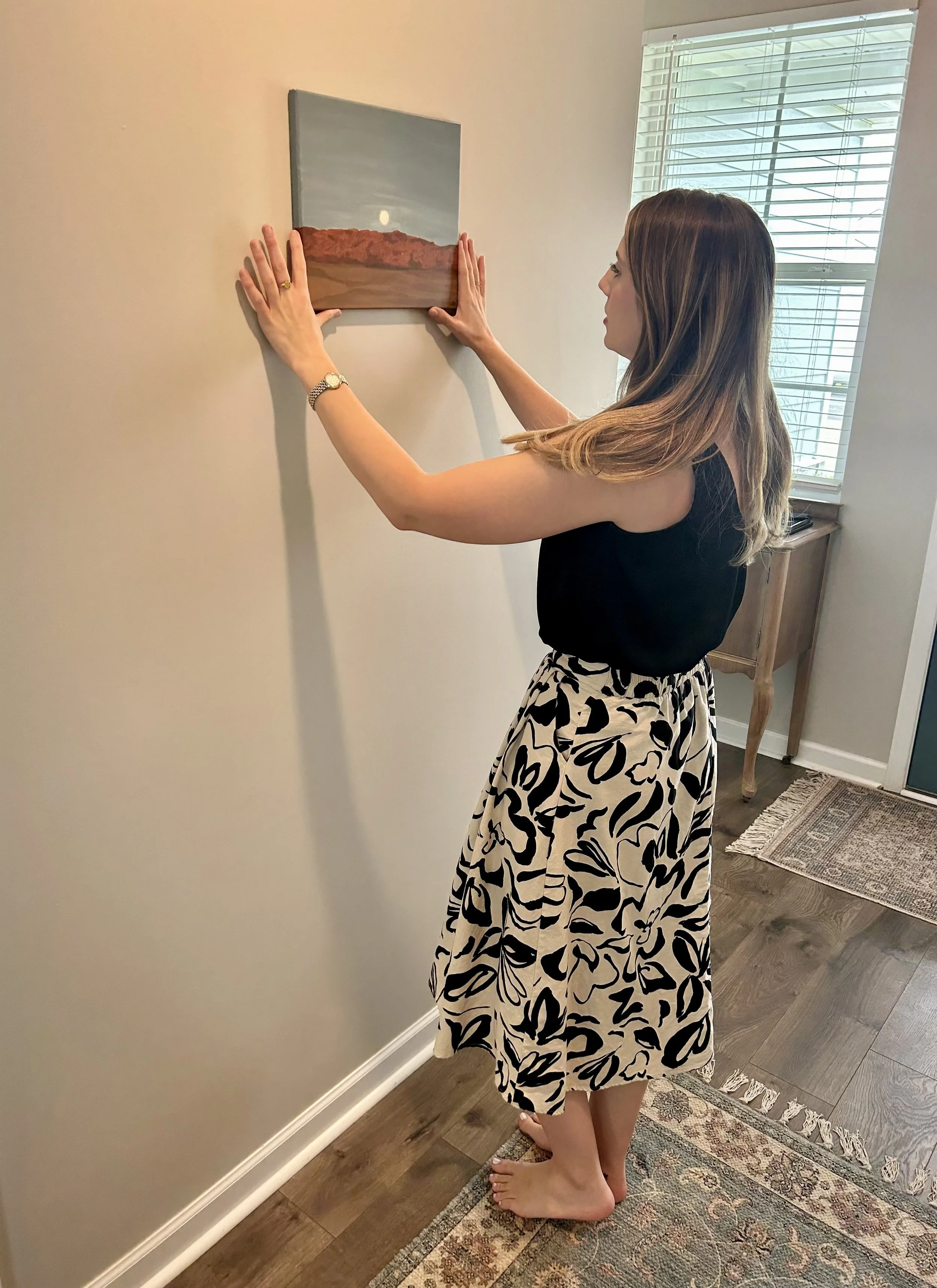 A woman with long brown hair, wearing a black sleeveless top and a black-and-white patterned skirt, is hanging a landscape painting on a beige wall in a well-lit room with wooden floor and a window with white blinds.
