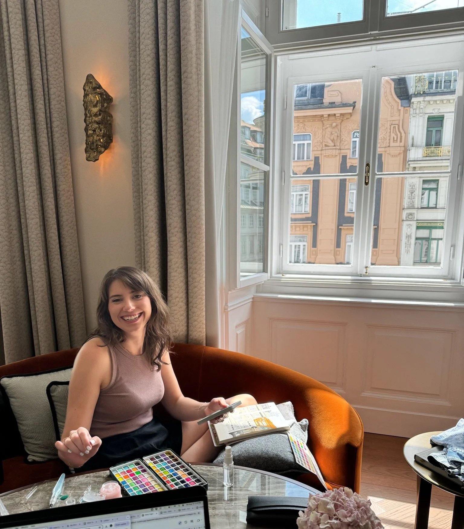 A woman with brown hair sitting on an orange velvet couch near a large window with city buildings outside. She is smiling and holding a makeup brush, with a makeup palette and other beauty supplies on the table in front of her.
