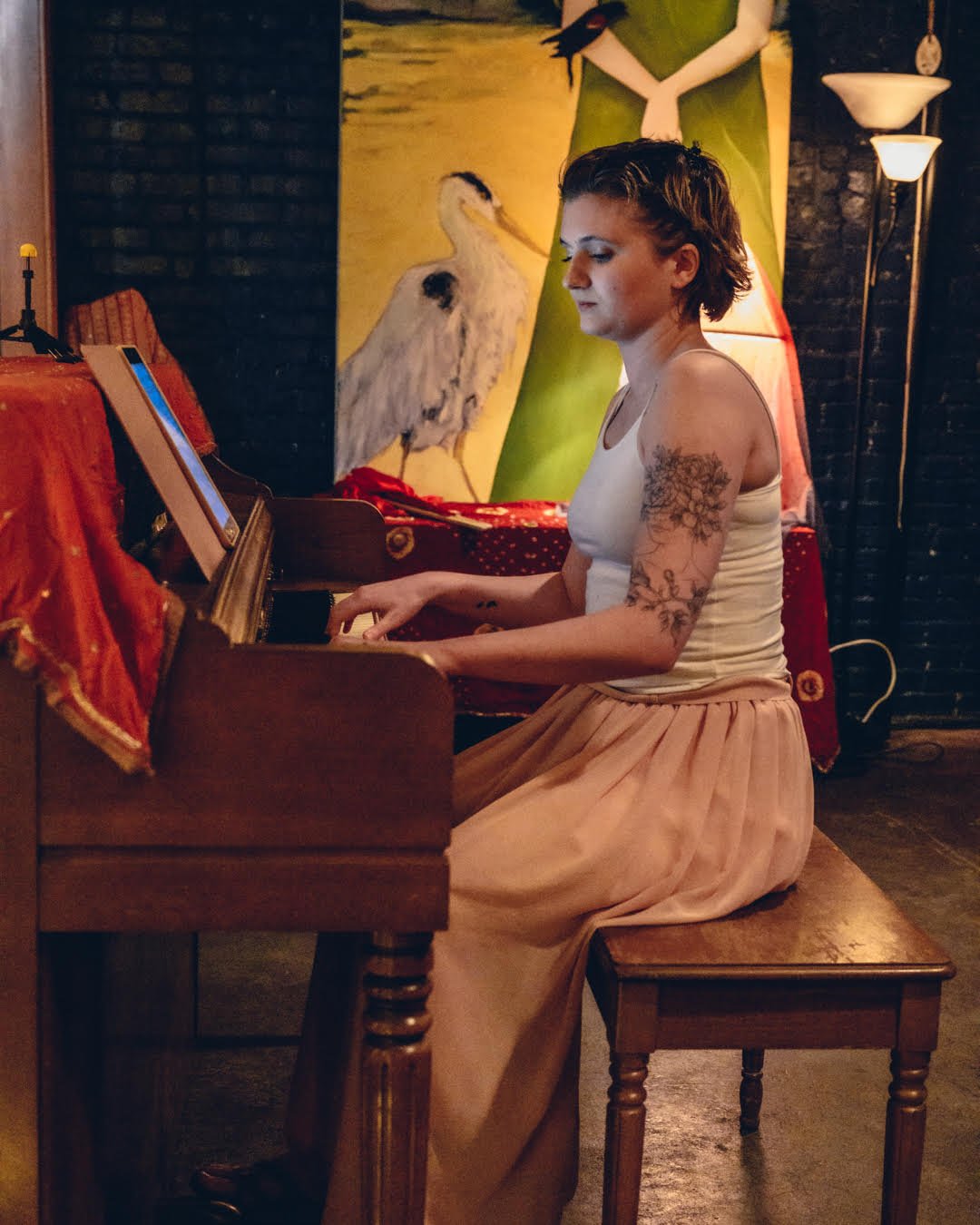 A woman with short hair and tattoos on her arms is sitting at a piano, playing. The setting appears to be a cozy, artsy room with warm lighting, a large colorful painting of a woman and a stork in the background, and red fabric covering part of the piano.