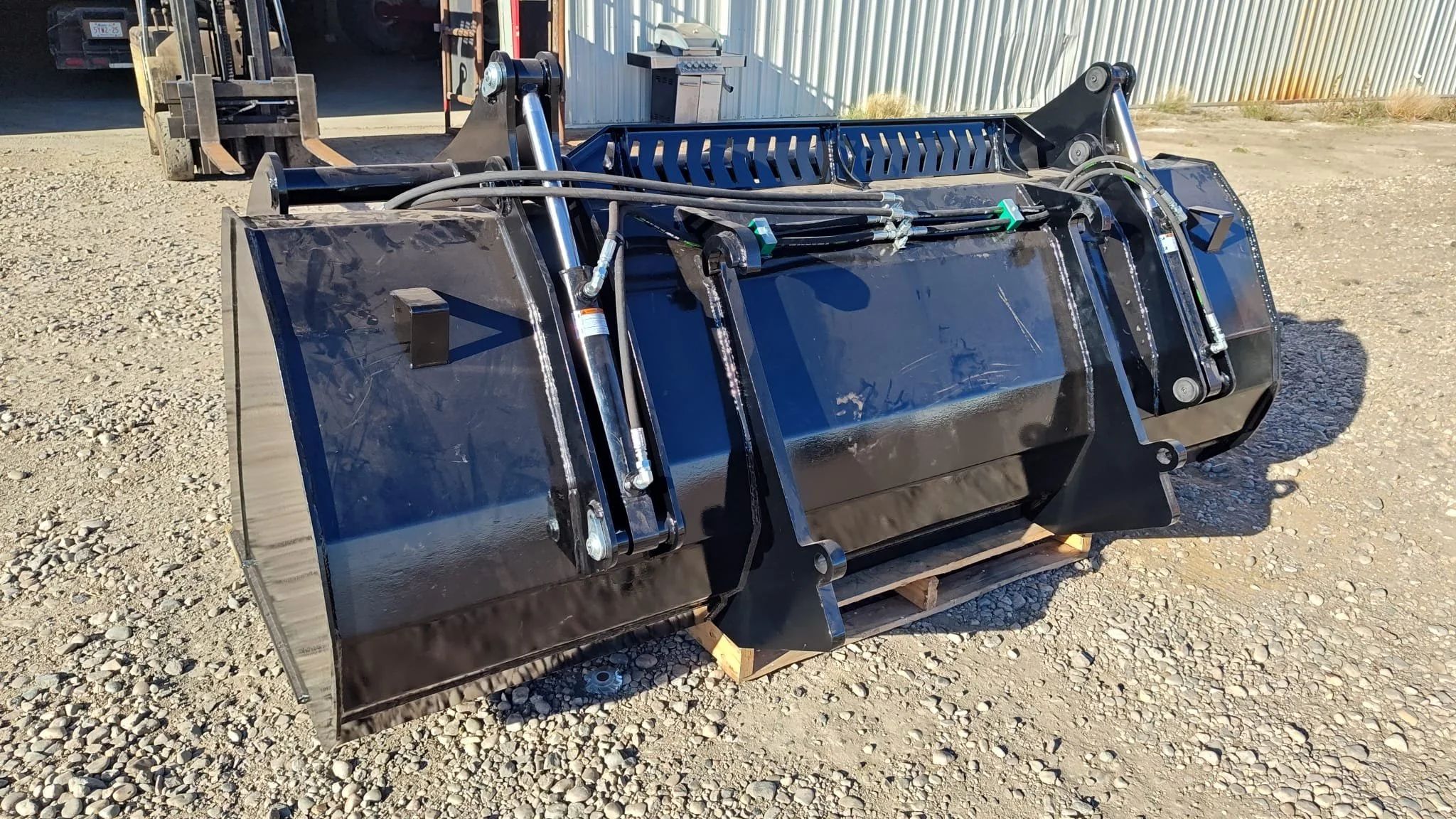 A large black industrial hydraulic attachment, likely for construction or excavation equipment, placed outdoors on gravel ground, with some equipment and a building in the background.