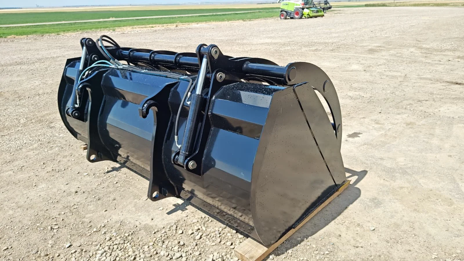 Close-up of a large, black, metal construction or agricultural implement attachment on a dirt field with green grass and tractor equipment in the background.