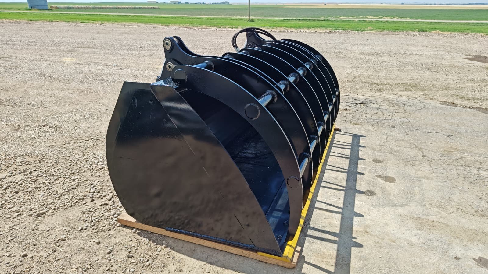 Large black industrial grapple bucket attachment placed outdoors on a concrete surface, with a rural landscape in the background.