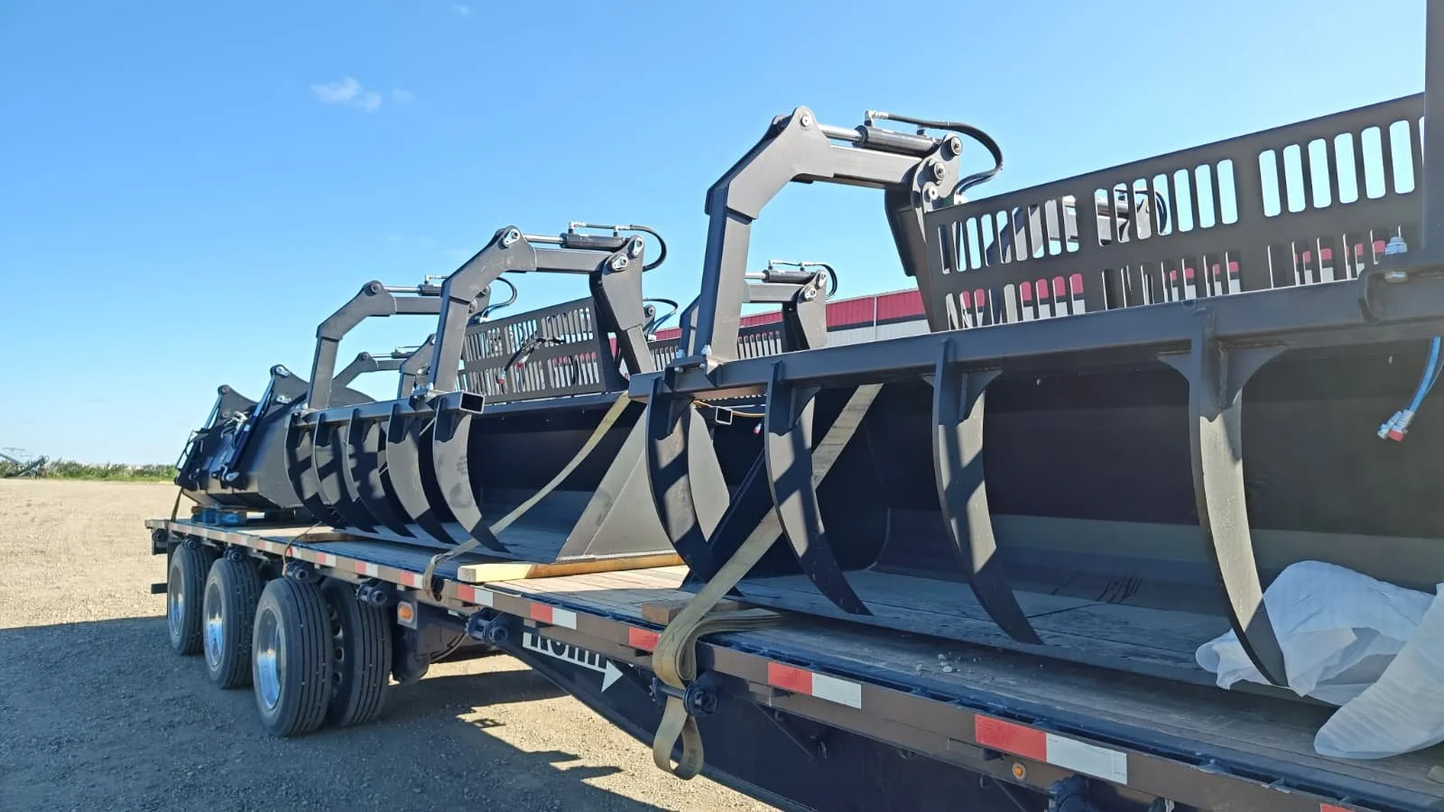 Multiple black excavator buckets loaded on a flatbed trailer outdoors under a blue sky.