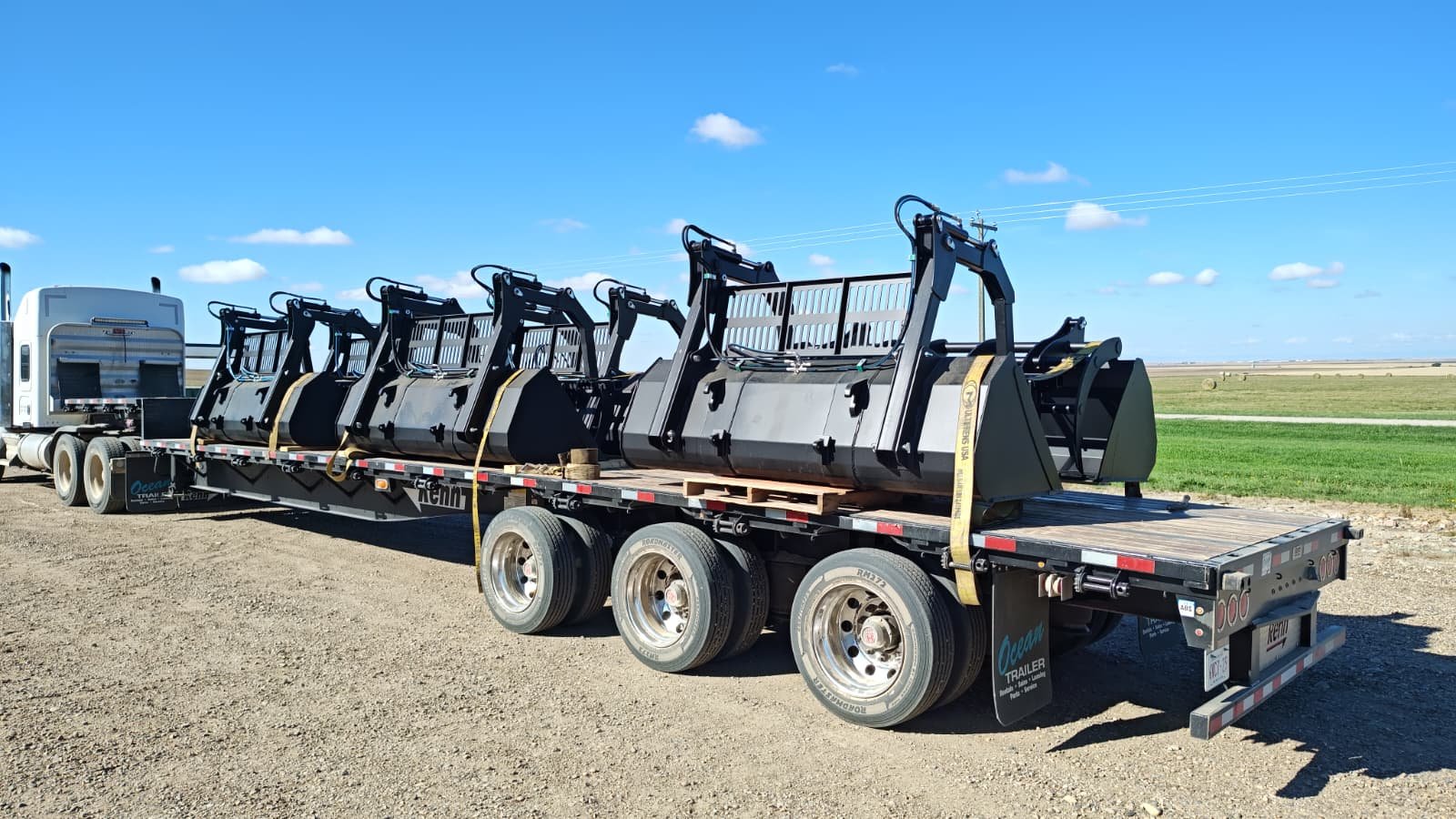 Flatbed truck carrying multiple large excavator buckets on a gravel lot with open fields and blue sky in the background.