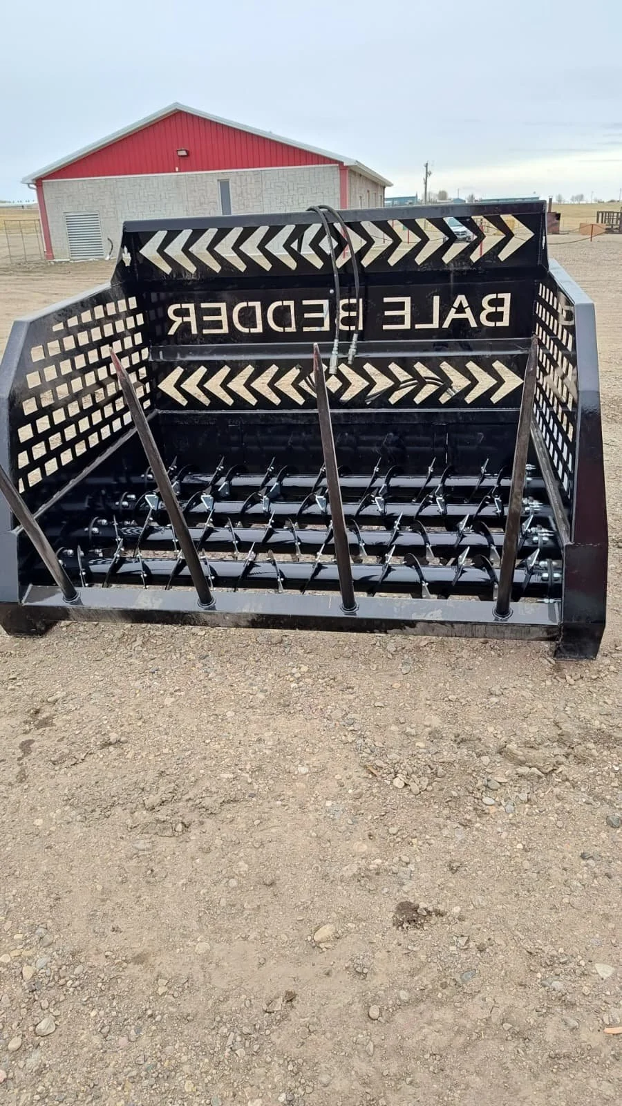 A black bale feeder for livestock on a gravel surface with a red barn in the background, under a cloudy sky.