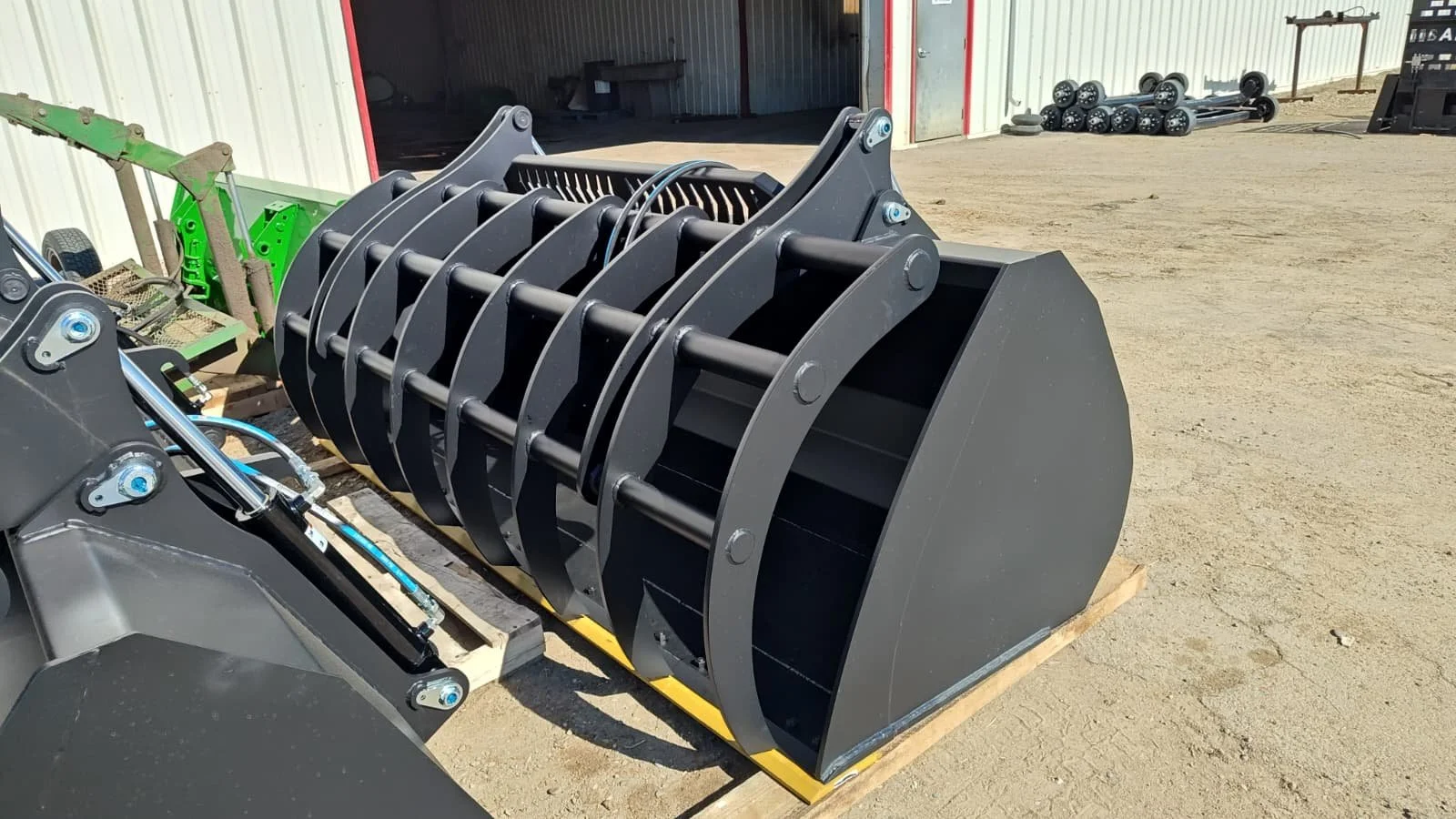 Large industrial grapple attachment for heavy equipment in an outdoor storage area, with a building and other equipment in the background.