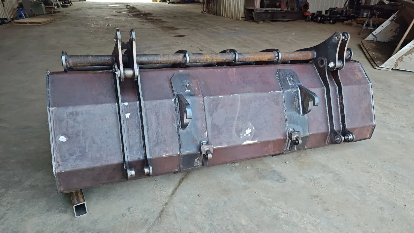 A large metal industrial bucket attachment with hydraulic arms resting on a concrete floor in a warehouse or workshop.