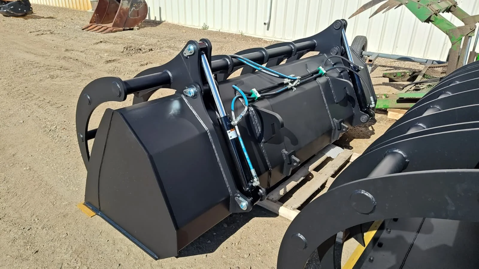 A large black industrial hydraulic implement with metal hydraulic cylinders and flexible hydraulic hoses, situated outdoors on a dirt surface, with other gardening or construction equipment in the background.