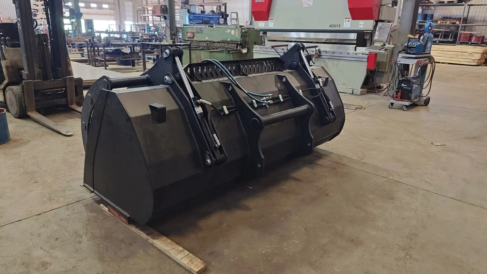 Industrial workshop with a large black mechanical bucket attachment for heavy equipment, surrounded by machinery, tools, and supplies.