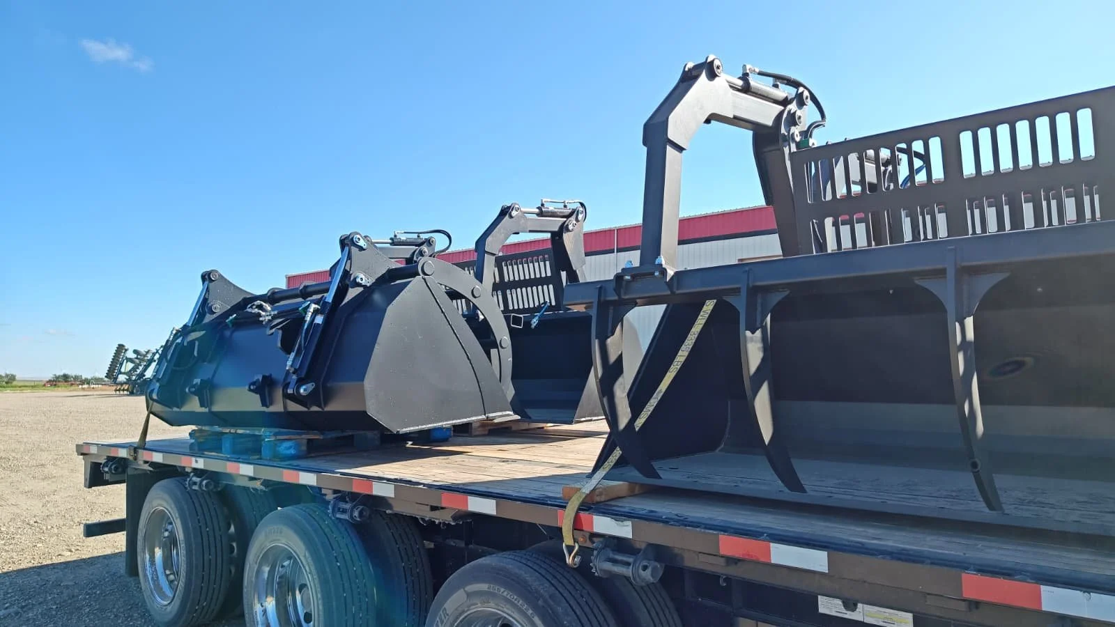 A flatbed truck carrying various heavy machinery parts, including a large black excavator bucket, located outdoors on a sunny day with a clear blue sky.
