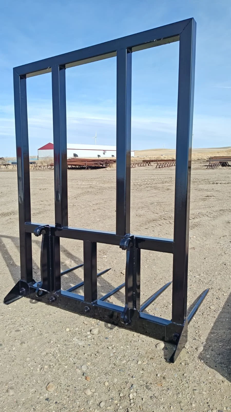 A black metal frame with four prongs at the bottom, set on a dirt ground outdoors, with a barn and pallets in the background under a blue sky.