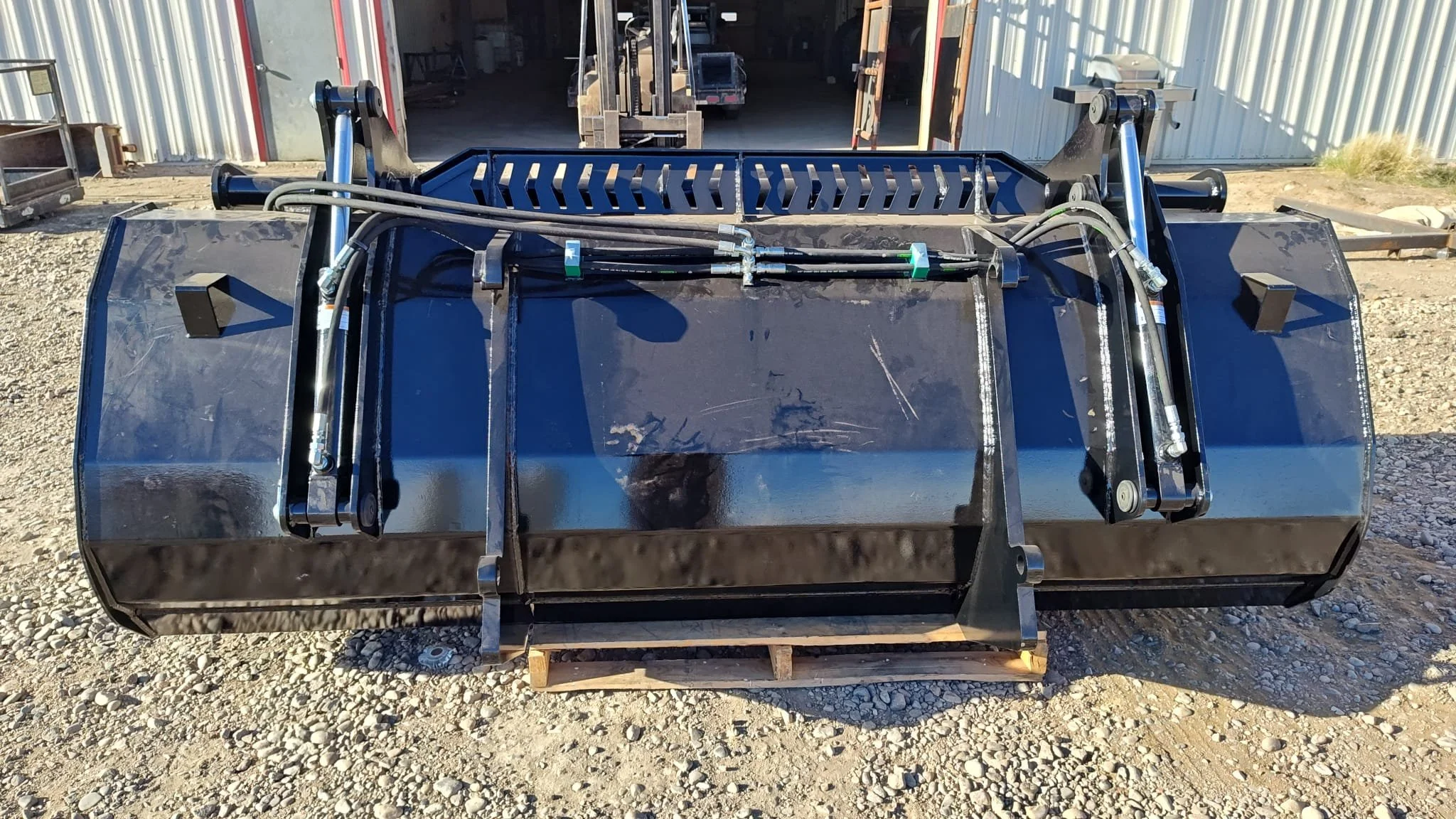 Front view of a large black industrial hydraulic attachment, possibly for construction equipment, with hydraulic hoses and metal components, positioned outdoors on a gravel surface near a metal building.