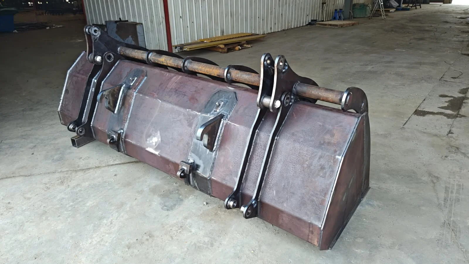 Industrial metal scraper with hinges and hydraulic cylinders on a concrete floor in a warehouse.