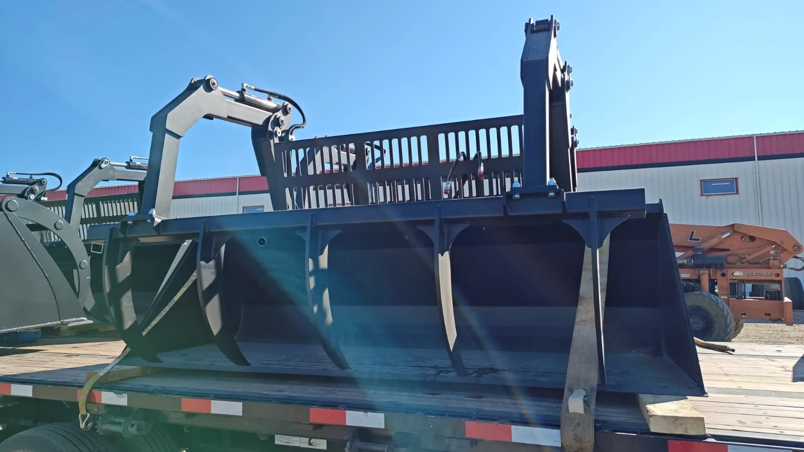 A large black industrial grapple attachment on a flatbed truck, used for handling logs or large materials, with machinery and a warehouse in the background under a clear blue sky.