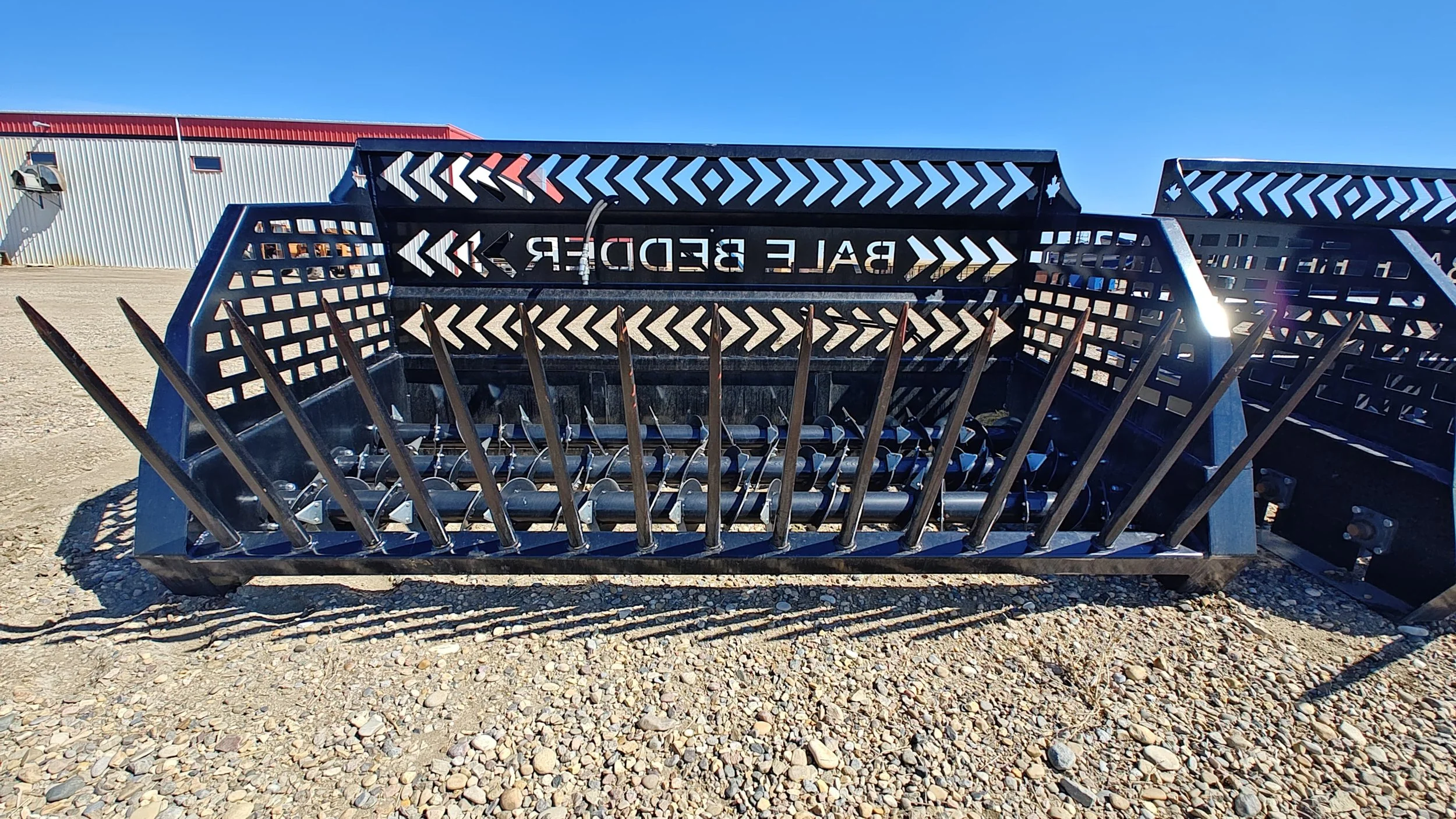 Metal agricultural equipment part with sharp spikes and a 'Bale Bedder' label, outdoors on gravel ground with a metal building in the background.