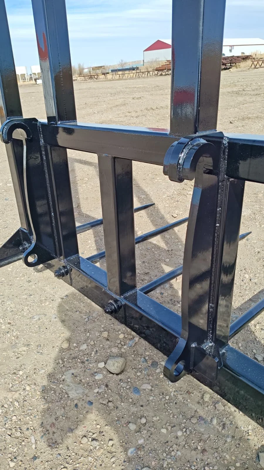 Close-up of a black metal cage or stand with hooks, set outdoors on dirt ground, with a barn and picnic tables in the background.