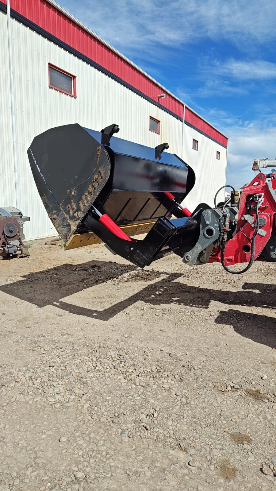 A black and red modern bucket attachment for a tractor, used for scooping and moving material, is raised off the ground in front of a large industrial building with a red and white exterior.