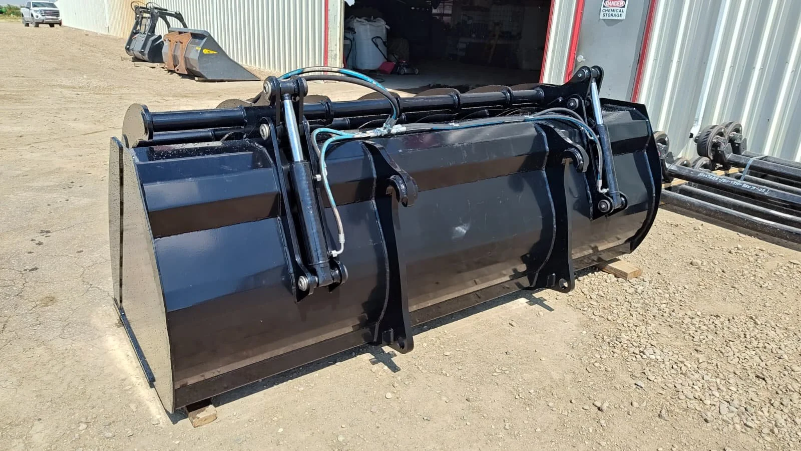 Large black industrial hydraulic attachment for construction machinery, resting on wooden blocks outside a warehouse