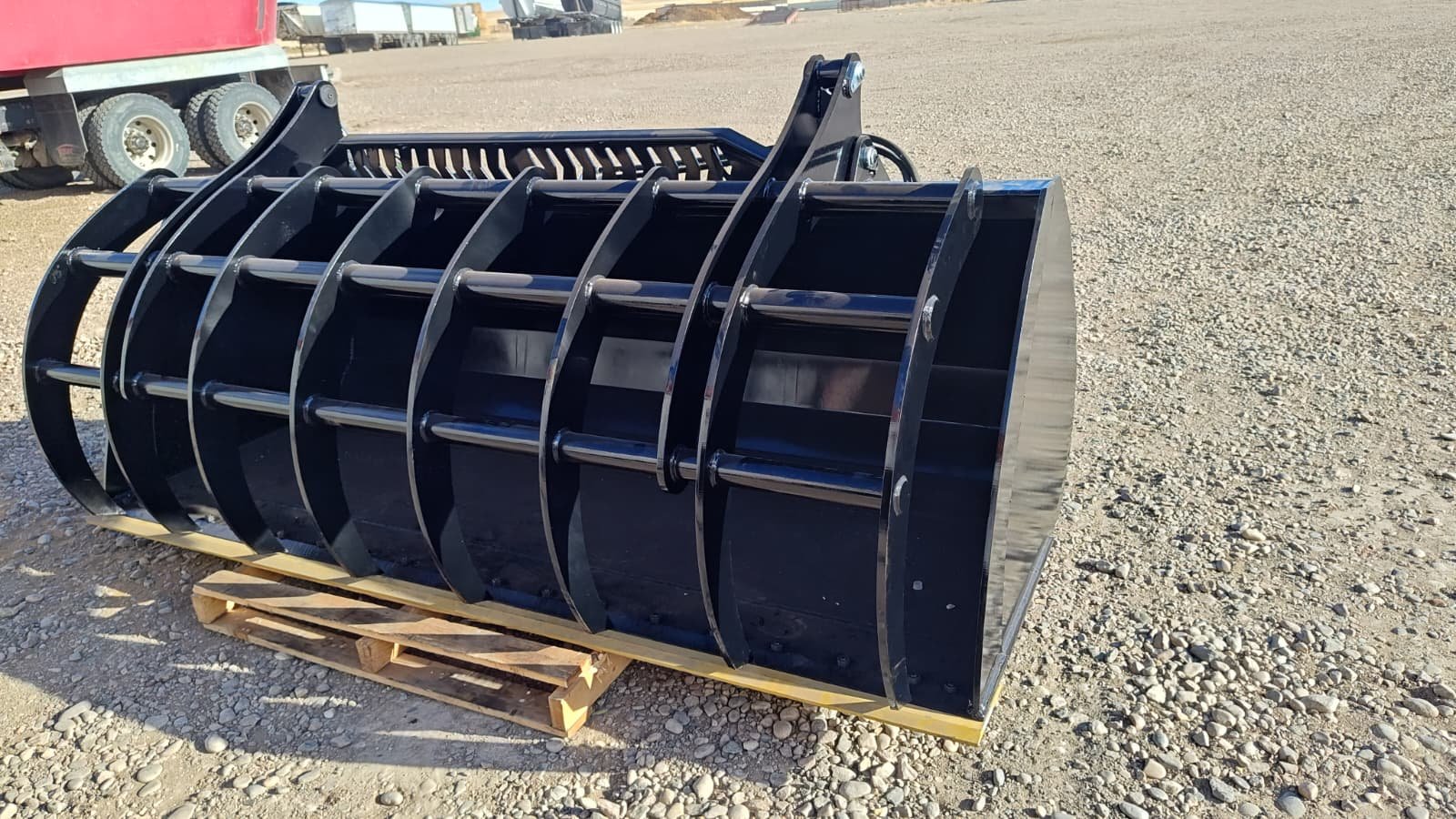 Large black industrial bucket attachment for heavy machinery resting on a wooden pallet on gravel ground.
