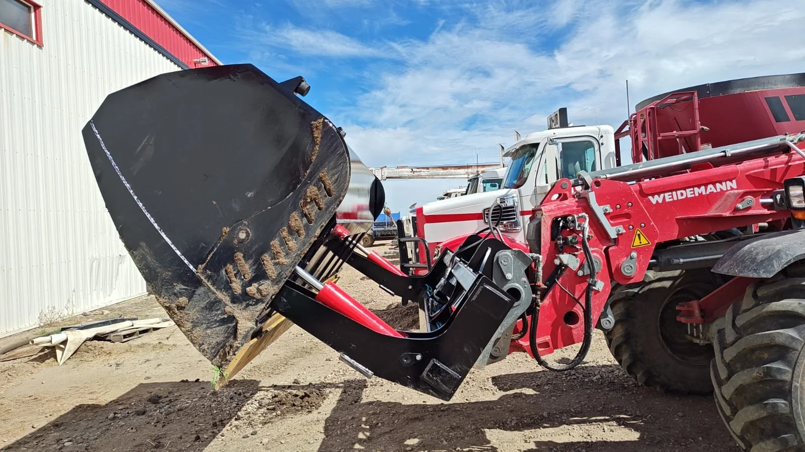 Close-up of a red and black agricultural tractor with a black plow attachment, parked outdoors on dirt ground, with a white building and blue sky in the background.
