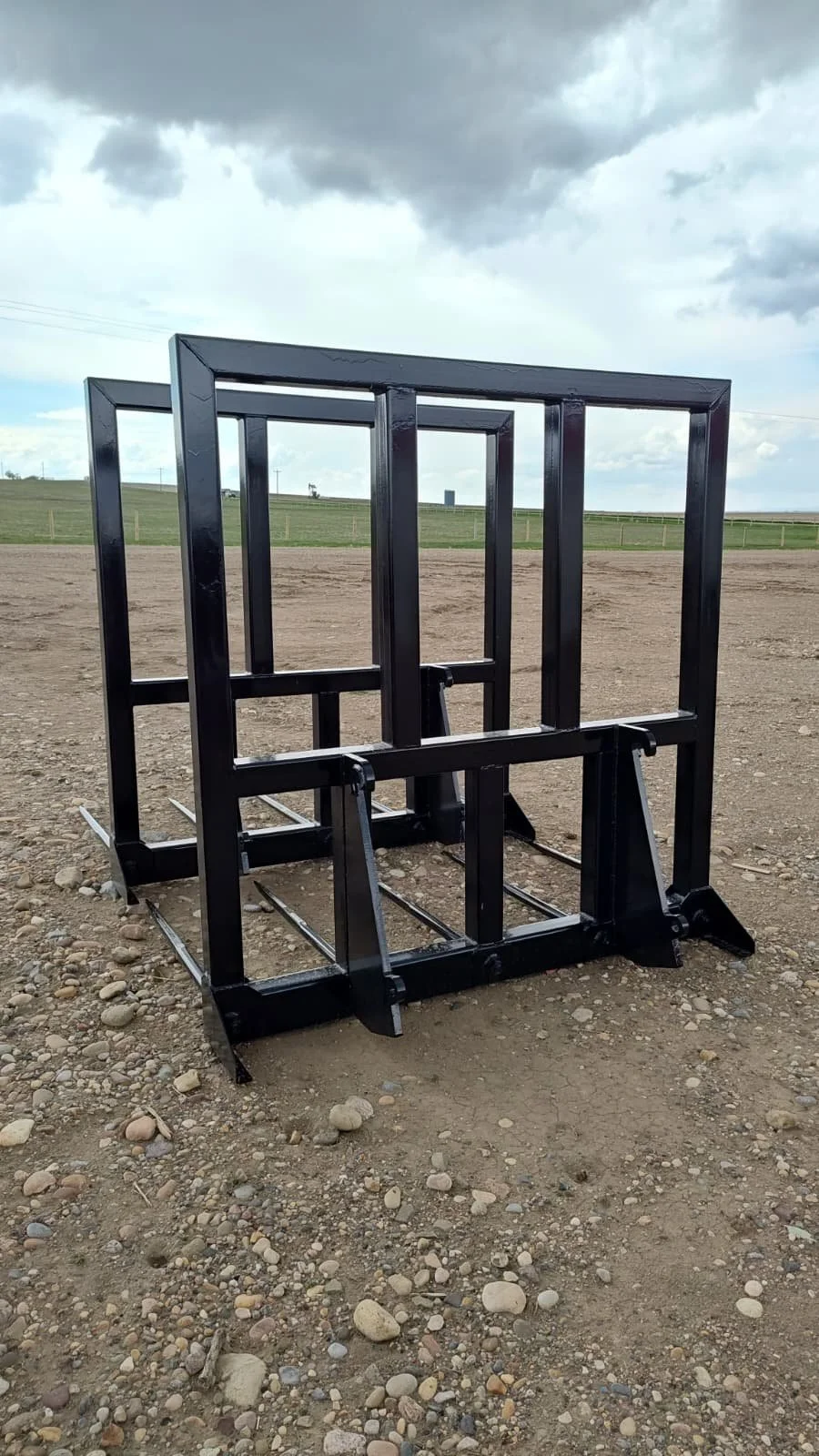 A black, metal sled with four runners and a rectangular frame, placed on dirt ground outdoors, under cloudy sky.