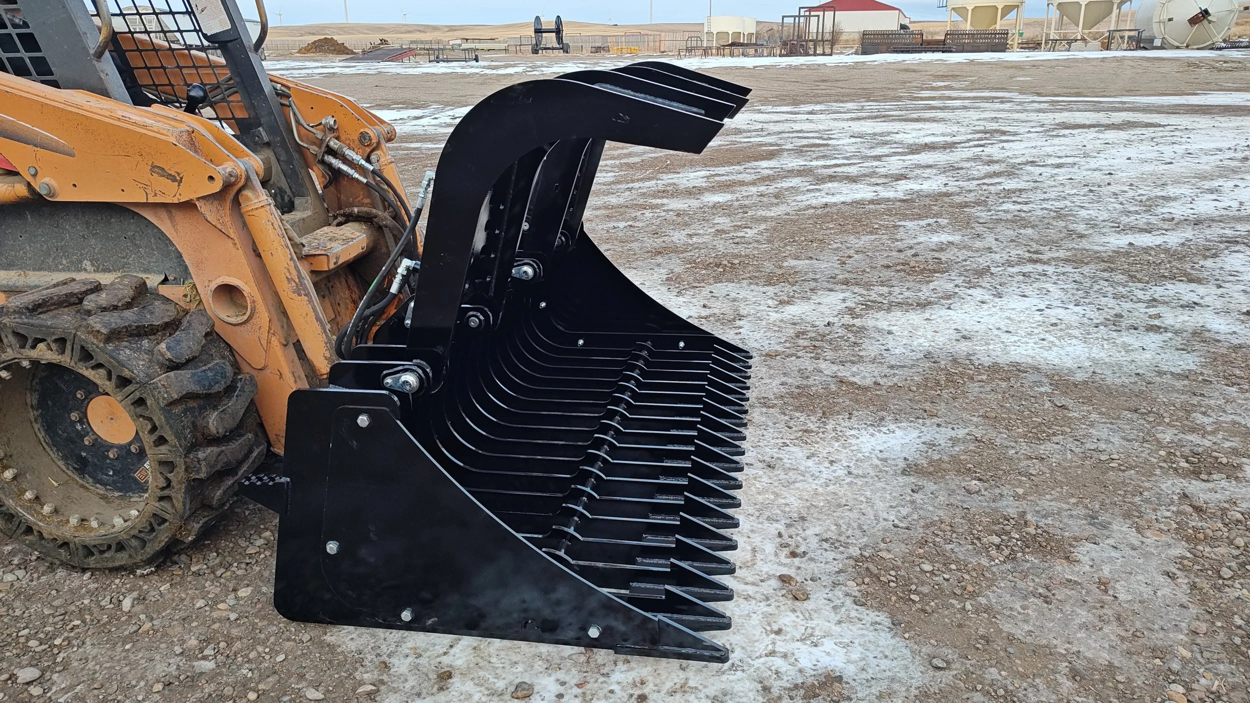 Close-up of a construction vehicle attachment with black metal blades on a muddy, partially snow-covered construction site with equipment and structures in the background.