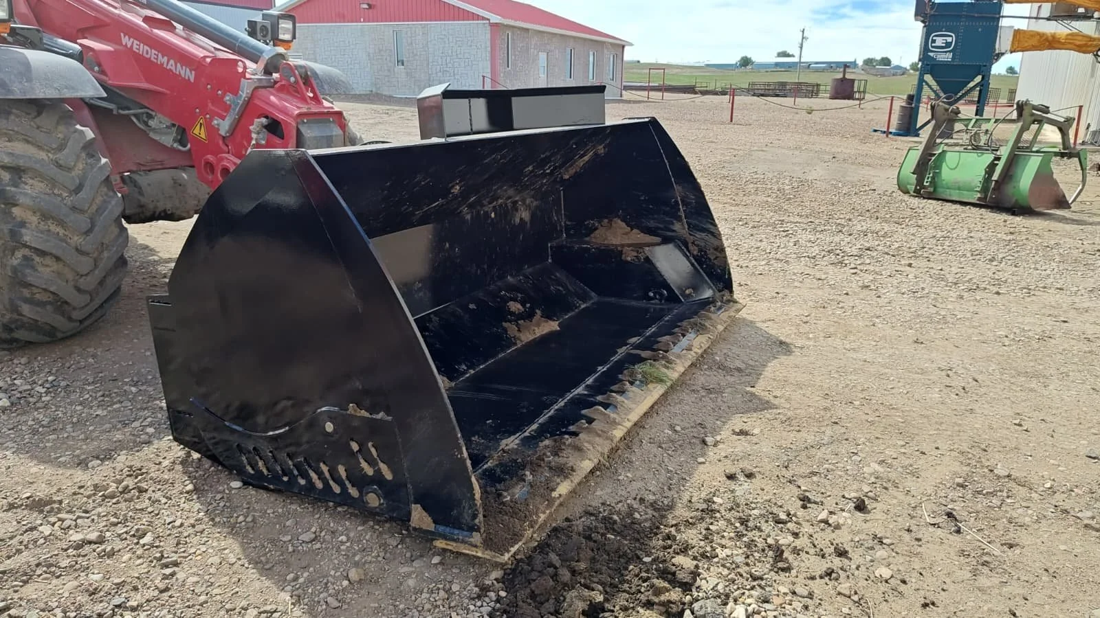 A large black bucket attachment for a construction or farming machine sitting on dirt ground, with parts of a red tractor and another construction vehicle visible in the background.