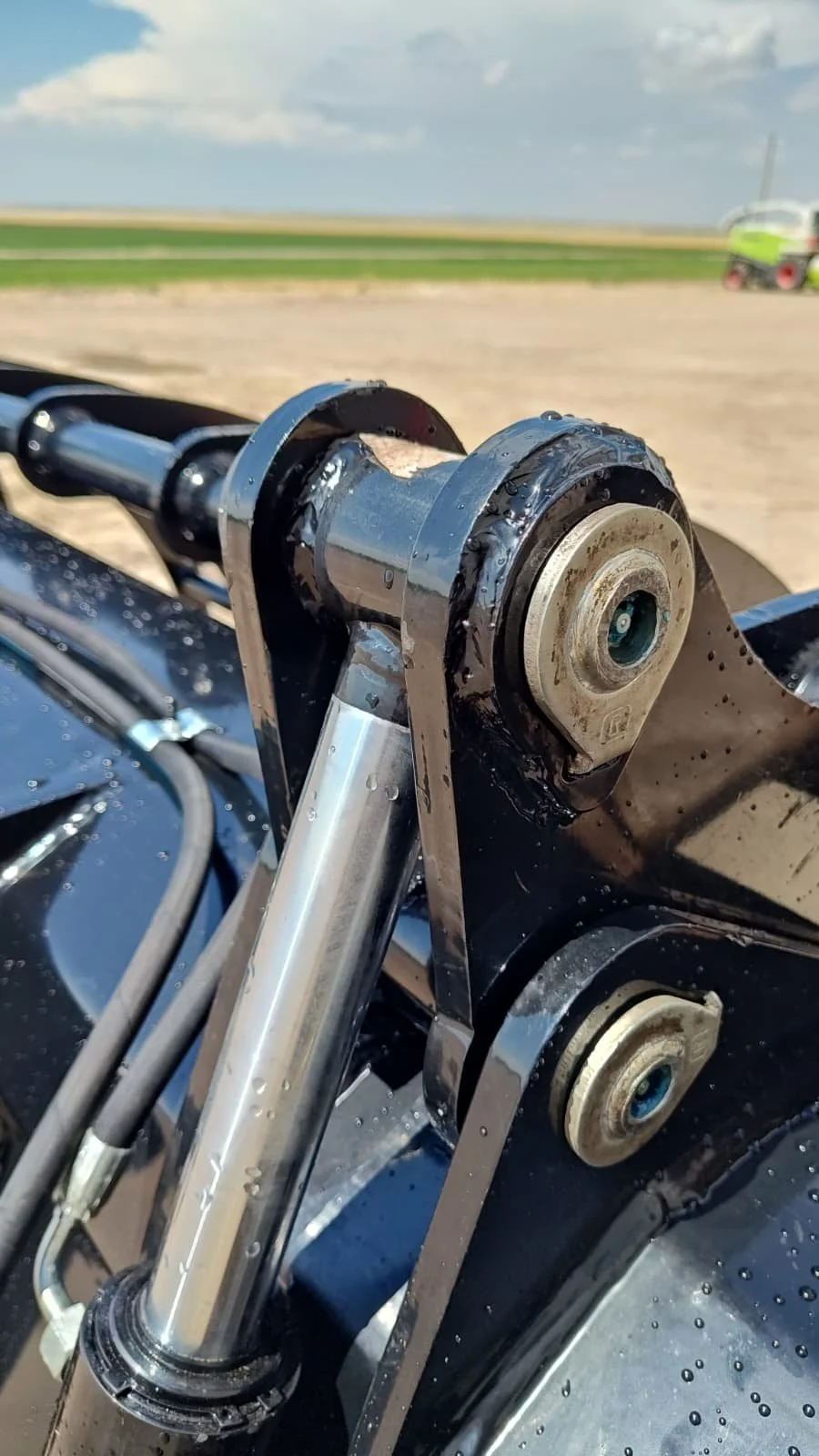 Close-up of a motorcycle front suspension and steering head with visible shiny fork tube, bearing, and frame mounting components, blurred landscape background.